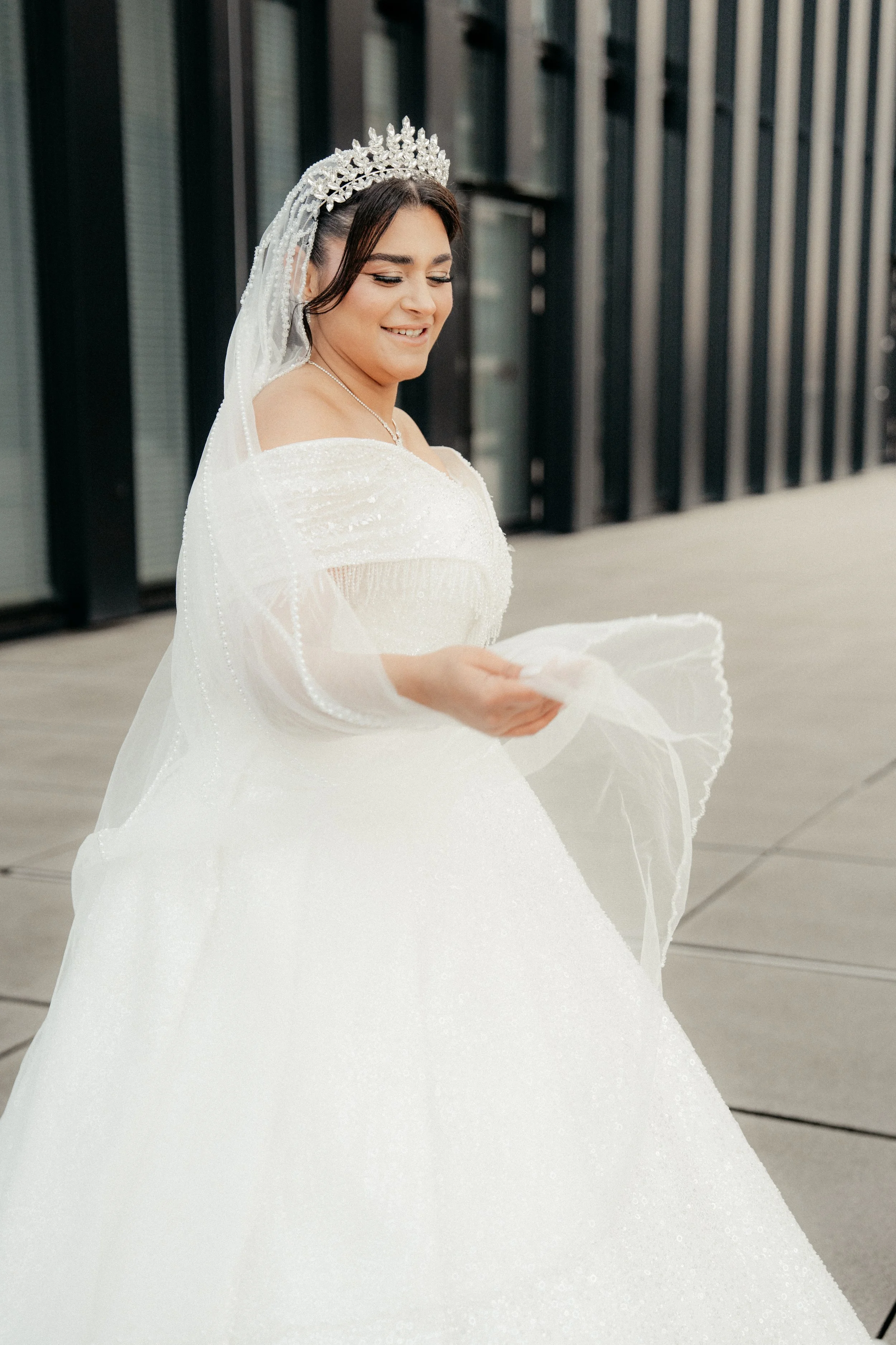 Braut in weißem Brautkleid mit Tiara und Schleier, lächelnd, im Freien vor einem modernen Gebäude mit schwarzen vertikalen Fassaden.