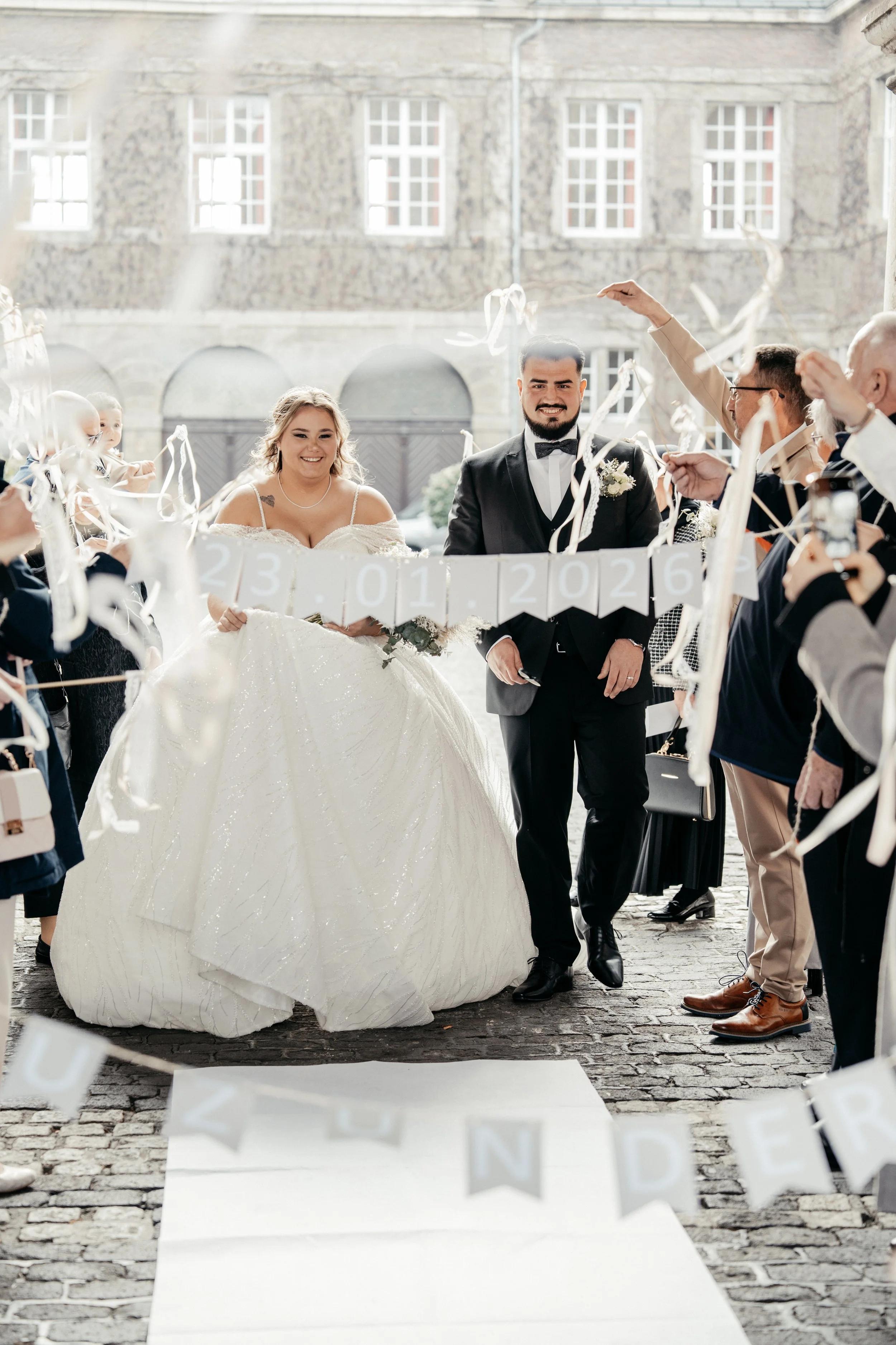Braut und Bräutigam feiern ihre Hochzeit im Freien, umgeben von Freunden und Familie, mit Dekorationen und bunten Bändern.
