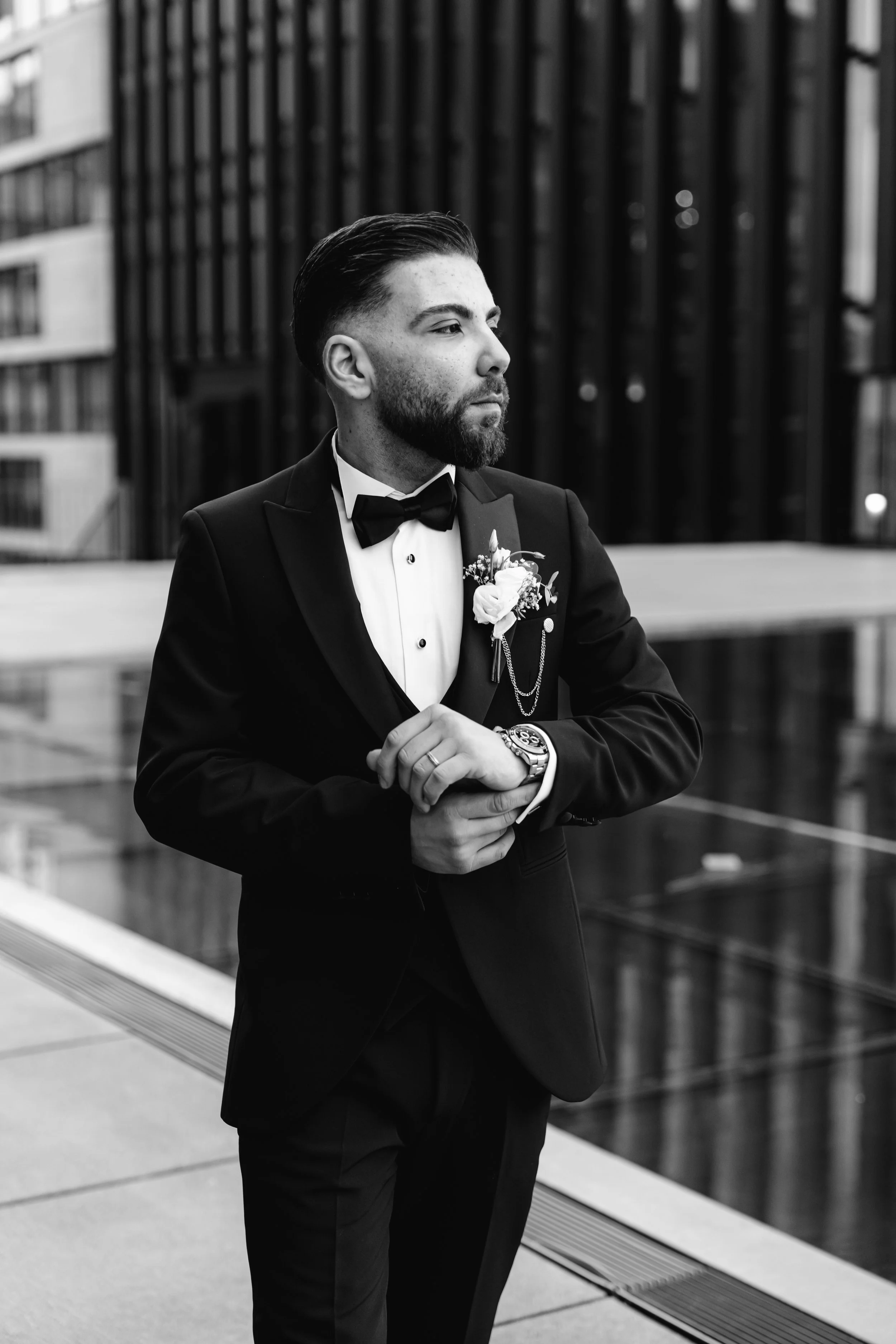 Ein Mann in einem Smoking steht in einer modernen urbanen Umgebung, schaut nach rechts, trägt eine Armbanduhr und hat ein Boutonnière am Revers auf der Brustseite.