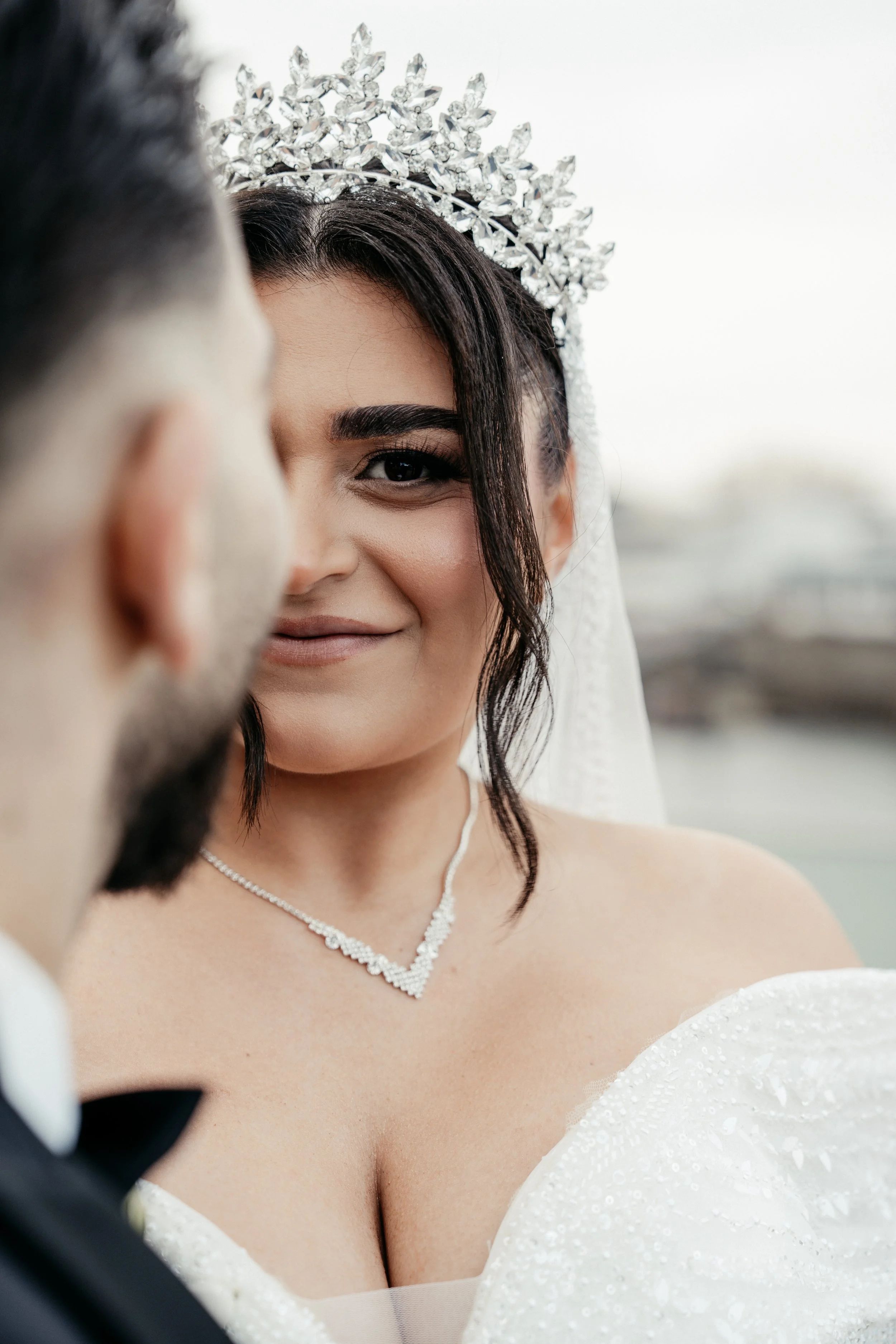 Ein Brautpaar bei einer Hochzeit, die Braut trägt ein weißes Kleid mit Perlen und eine funkelnde Tiara, während sie sich gegenseitig anschauen.