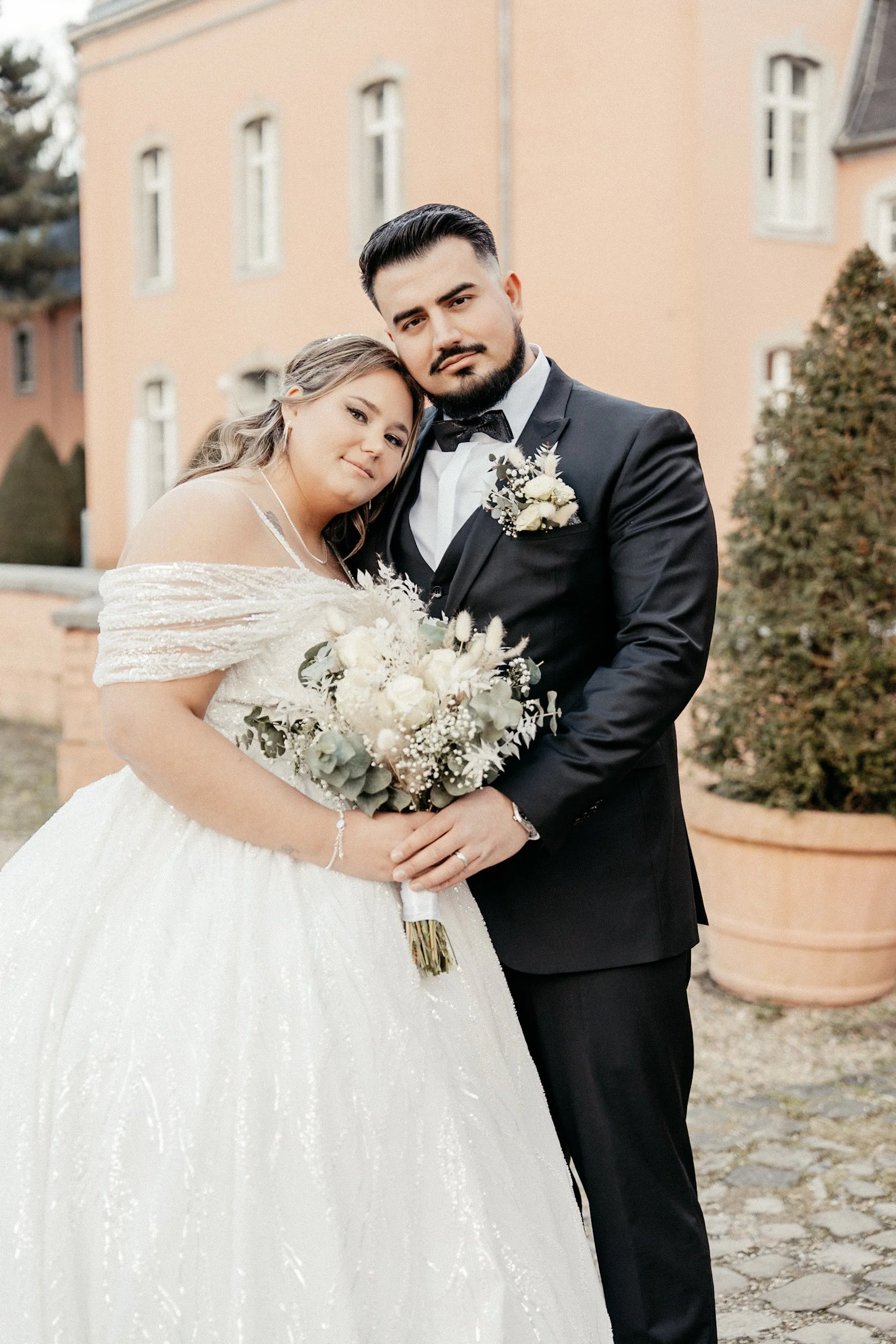 Ein Brautpaar bei einer Hochzeitsaufnahme, die Braut trägt ein weißes Kleid und hält einen Blumenstrauß, der Bräutigam trägt einen schwarzen Anzug und eine Fliege.
