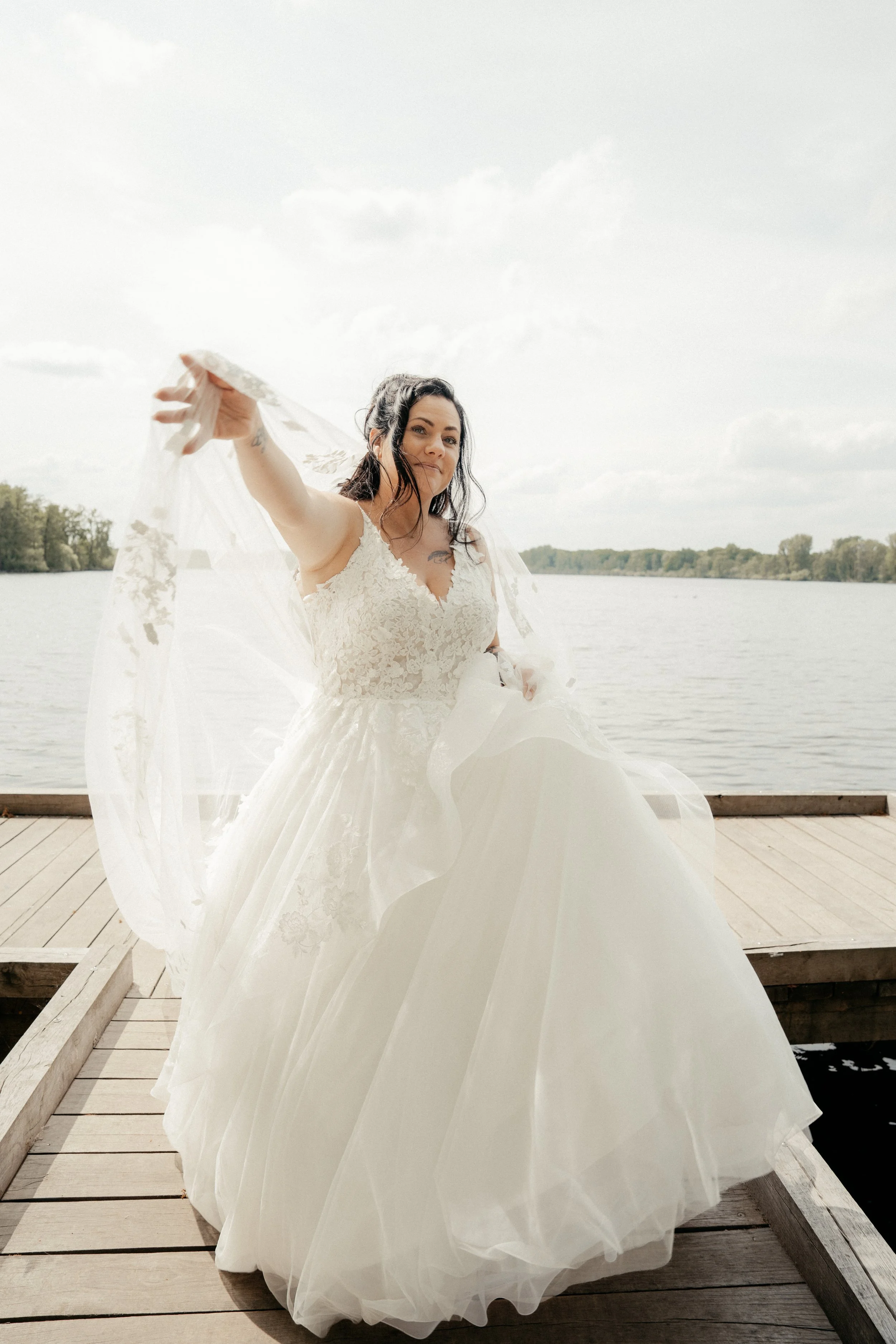 Eine Braut in einem weißen Hochzeitskleid, die auf einem Holzsteg am Wasser steht, mit einem ländlichen Hintergrund aus Bäumen bei bewölktem Himmel.