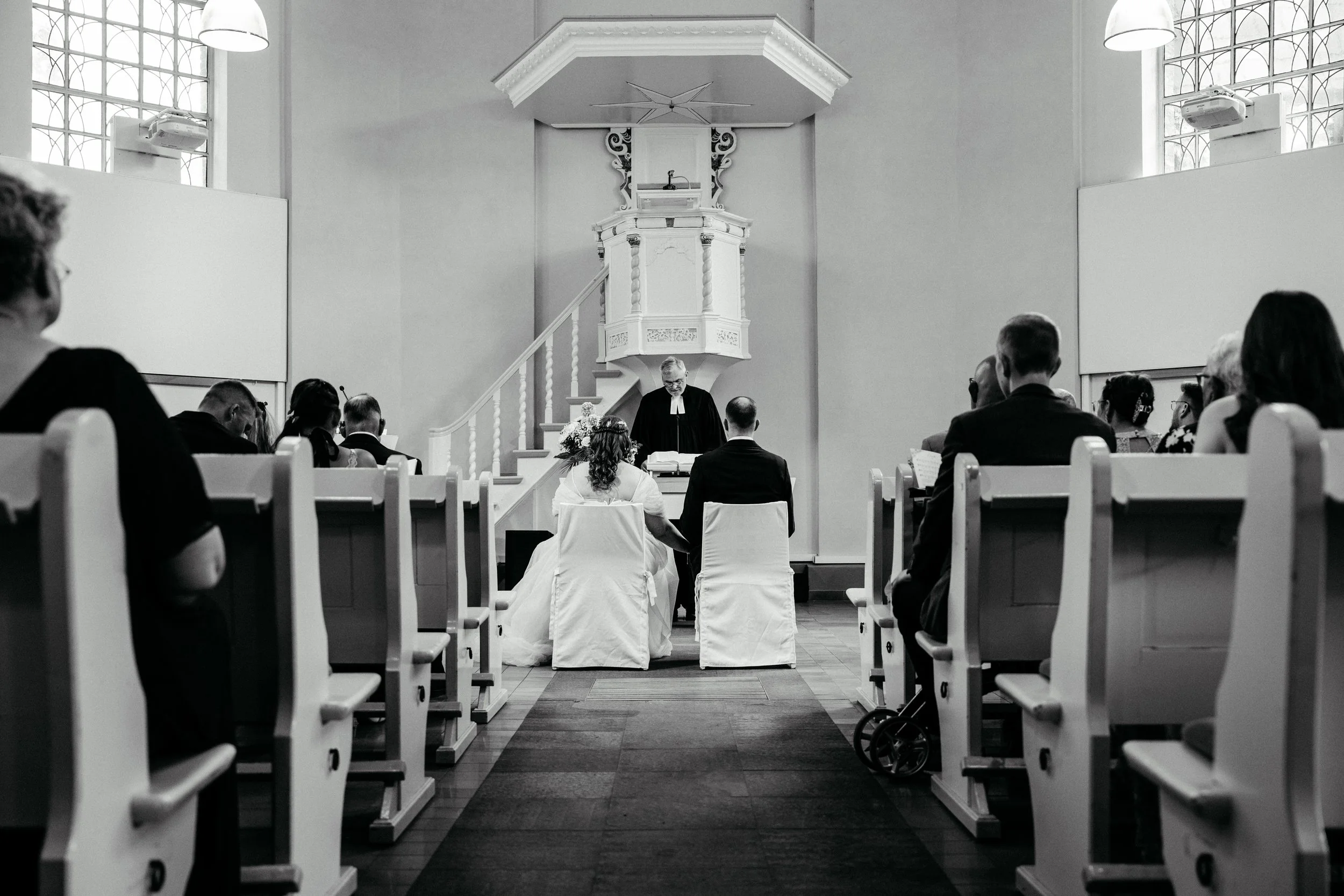 Schwarz-Weiß-Foto einer Trauung in einer Kirche mit vielen Gästen. Das Brautpaar steht vor dem Pfarrer an einem Altar, im Hintergrund große Fenster und eine Kanzel.