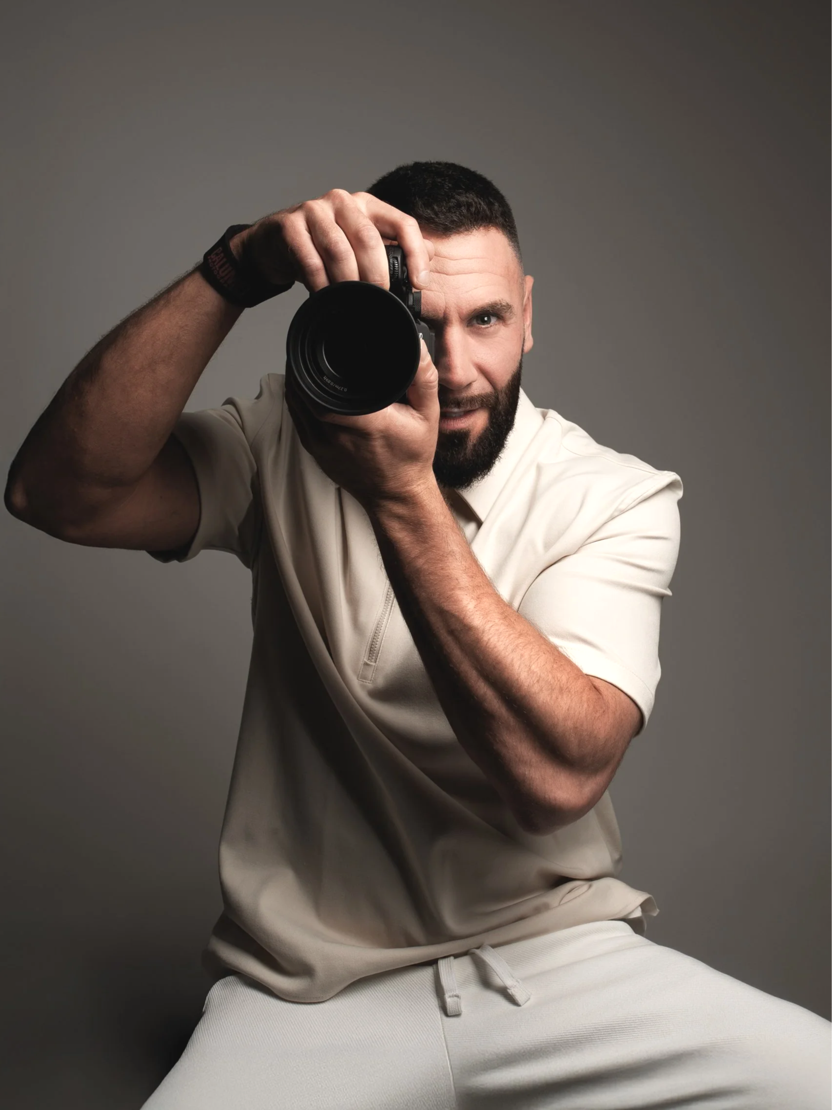 Mann in weißen Sportkleidung hält eine Kamera und fotografiert direkt in die Kamera. Er sitzt vor einem grauen Hintergrund.