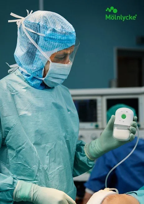 A healthcare worker in scrubs, mask, and gloves using a medical device in a hospital setting.