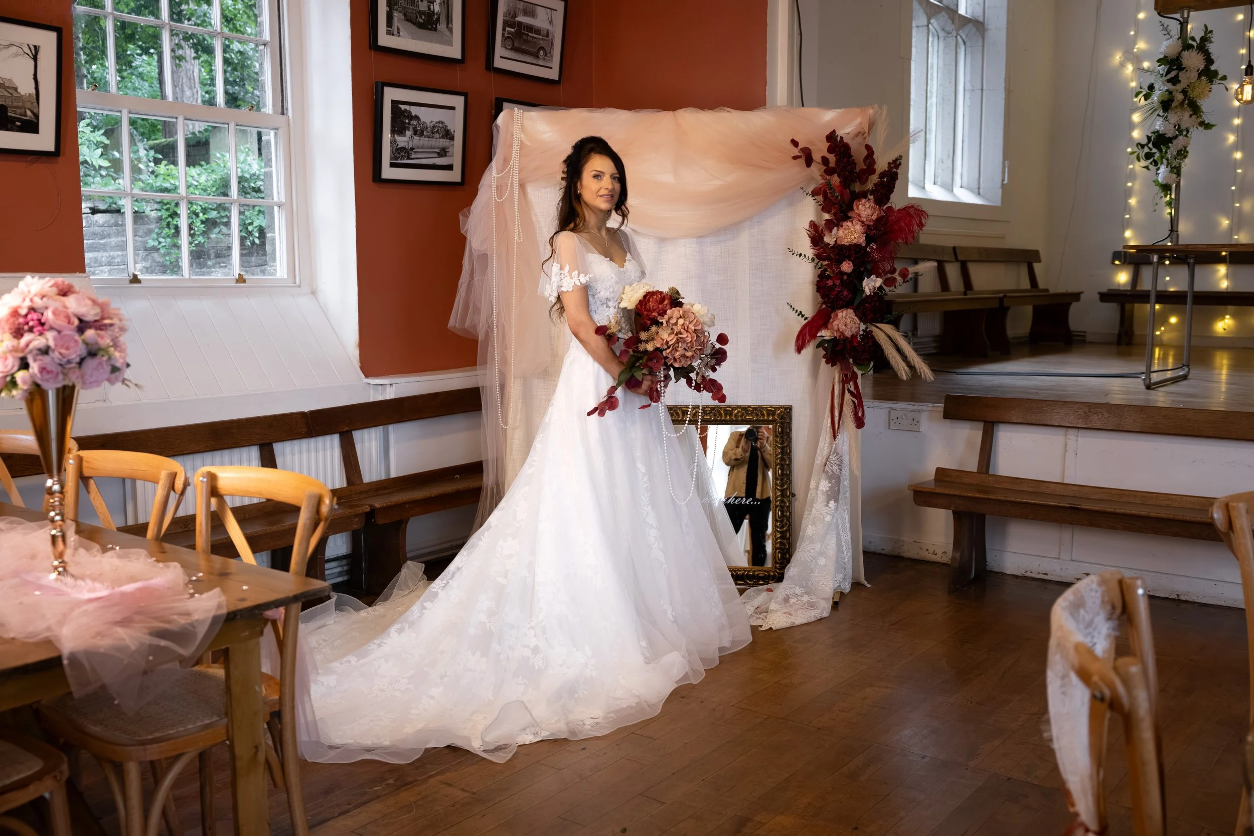 A woman in a wedding dress holding a bouquet, posing indoors near floral decorations and a mirror, with photographs on the wall and a window showing greenery outside. Inside wedding venue the Old School Rooms Haworth