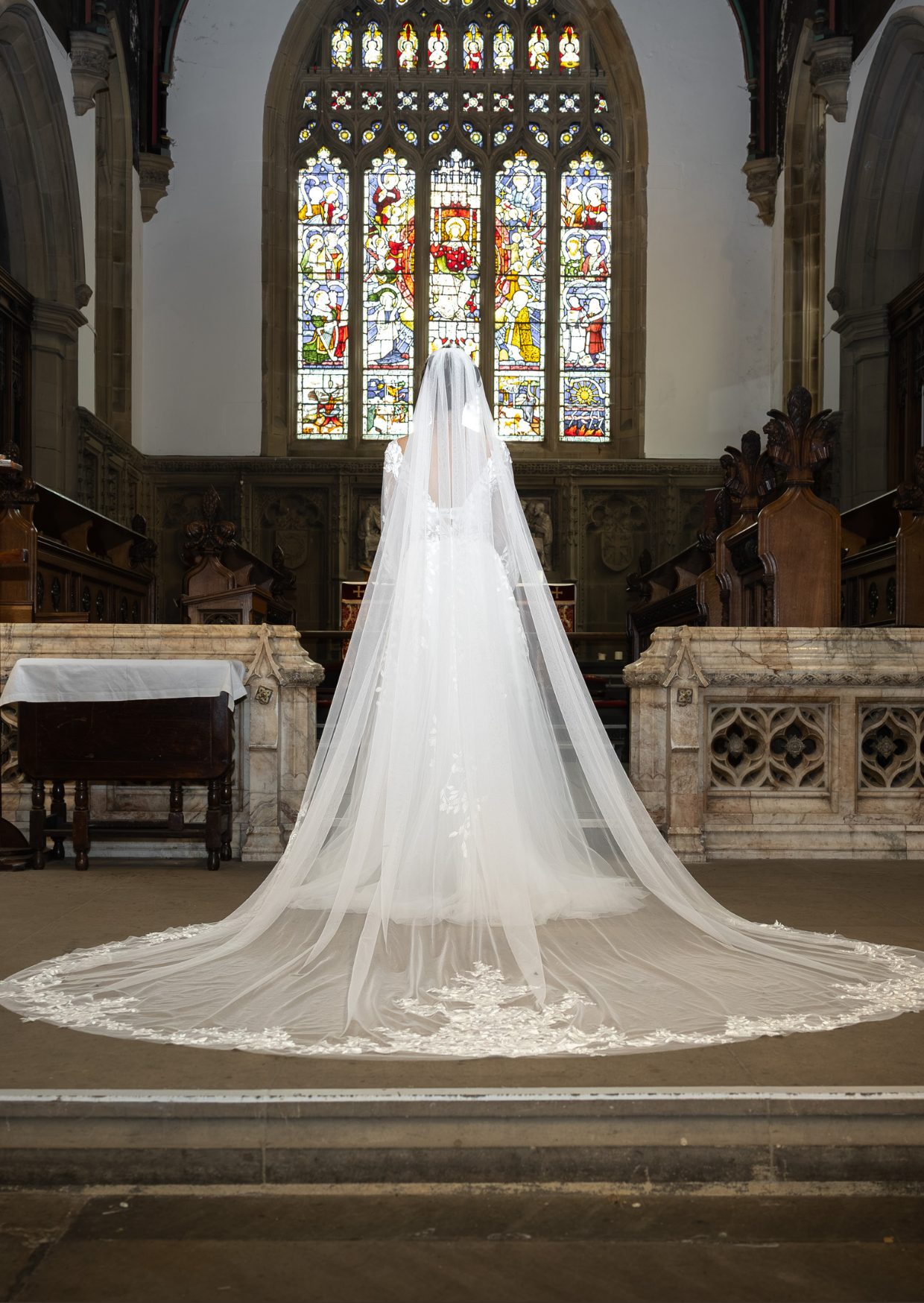 A bride in a white wedding dress with a long train and veil standing inside Haworth church, facing a large stained glass window that depicts religious figures and scenes.
