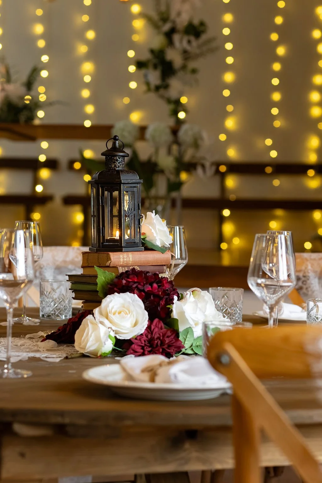 Decorated dining table with a black lantern, stacked books, white and red flowers, and glassware, with yellow fairy lights in the background. A Bronte Sister inspired theme