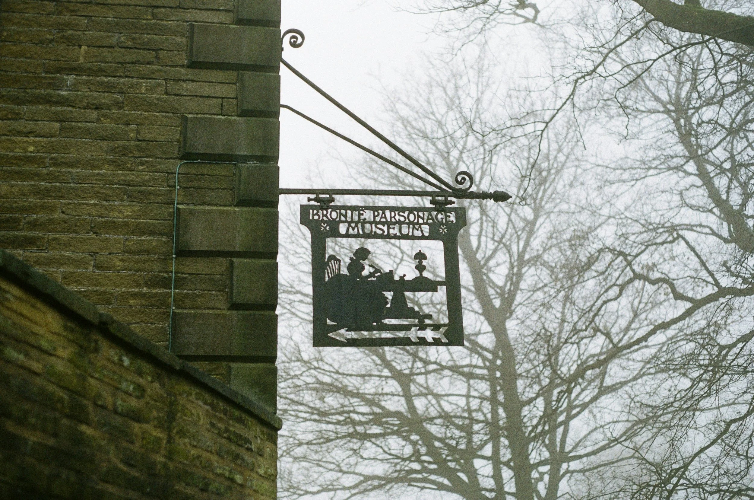A black metal sign for the Bronte Parsonage Museum hanging from a brick building on a foggy day, with bare tree branches in the background.