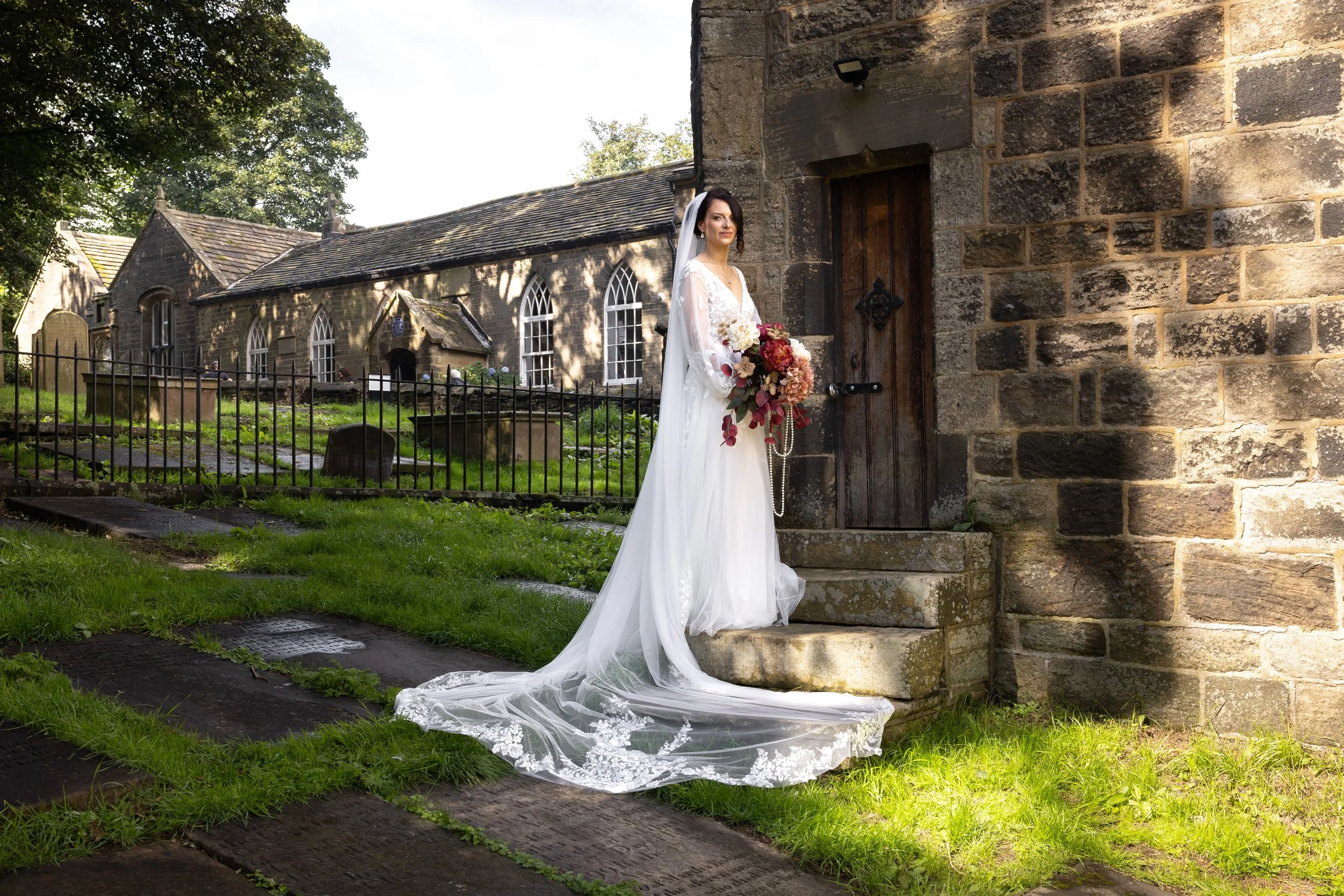 A bride in a white wedding gown with long train and lace details, holding a bouquet of pink, white, and peach flowers, standing outside a stone church in Haworth with gothic windows, against a background of trees and a cloudy sky.