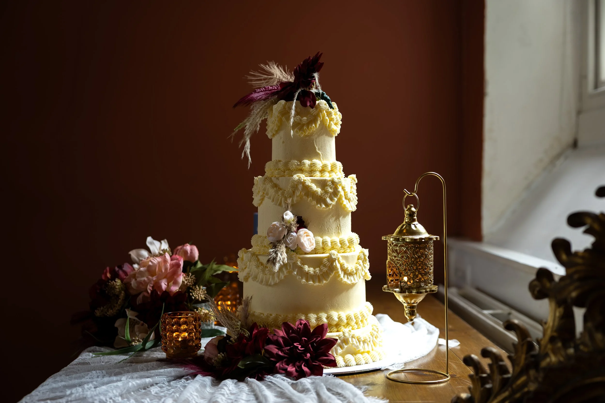 A tall, multi-tiered cream-colored wedding cake decorated with cream piping, flowers, and botanical accents, situated on a table with floral arrangements and a lace tablecloth, near a window with sunlight. In the Old School Room, Haworth