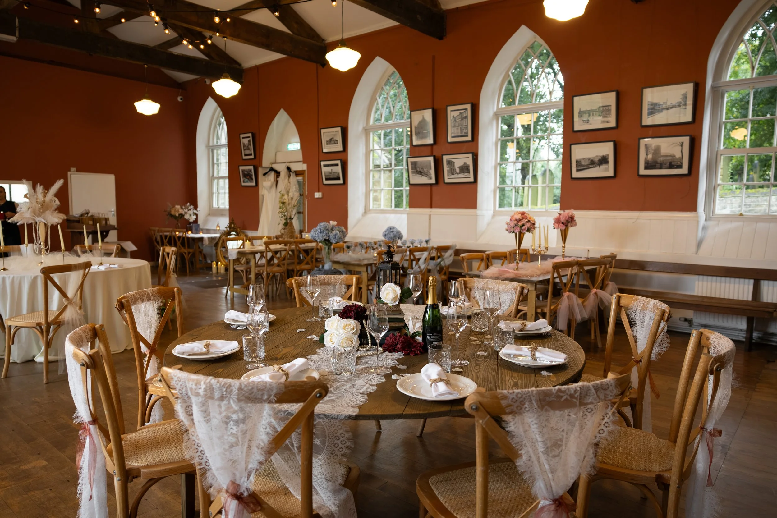 Indoor banquet hall decorated for a wedding reception with round tables covered in lace tablecloths, floral centerpieces, wine glasses, and wrapped chairs. The Old School Rooms, Haworth