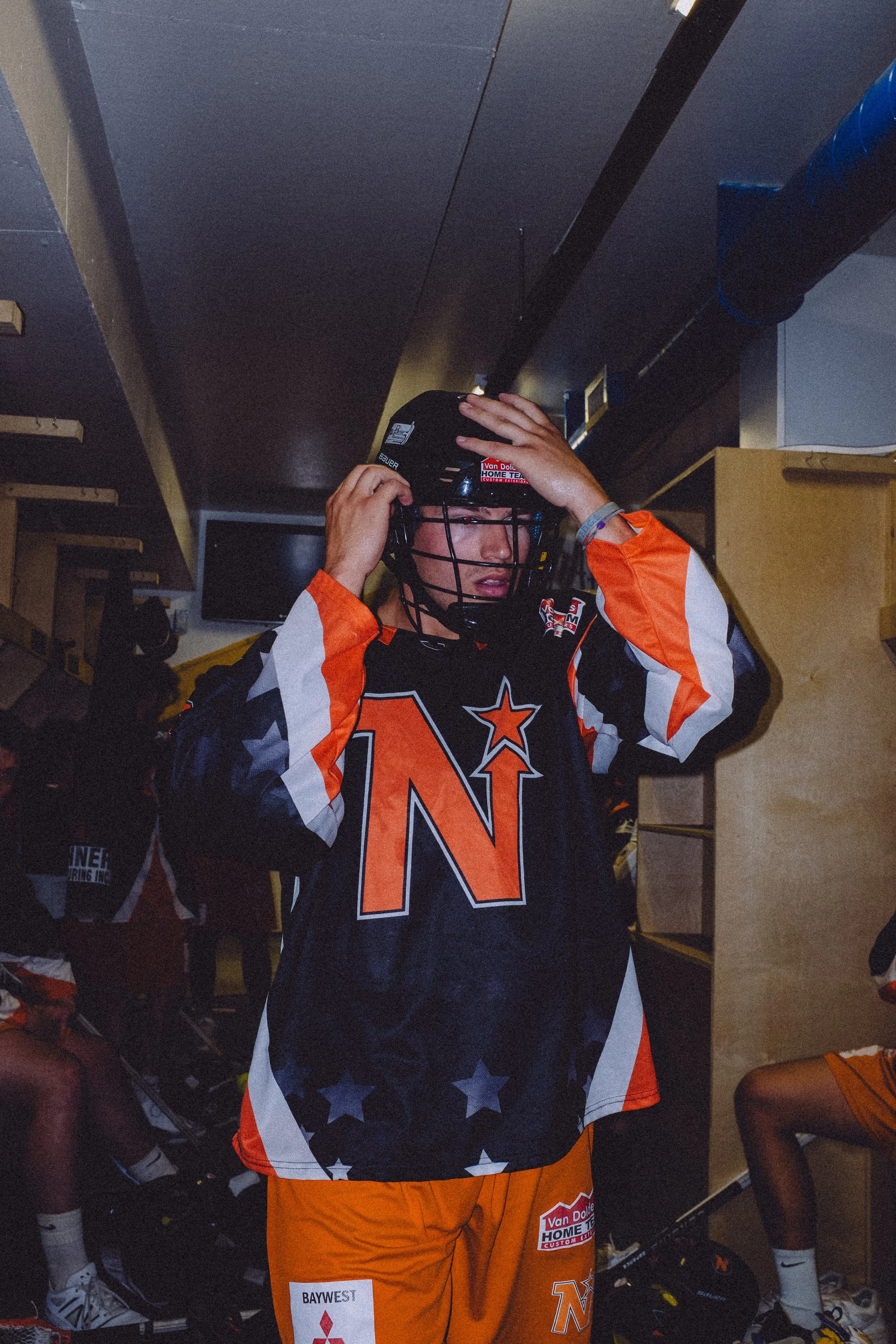A box lacrosse player putting on a black helmet in a locker room, wearing a black and orange jersey with stars and the Owen Sound North Stars logo on it, and orange shorts.