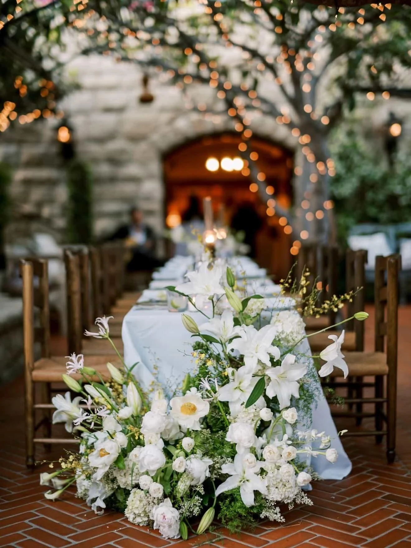 A long rectangular table covered with a white tablecloth, decorated with a large arrangement of white lilies, roses, and other flowers, set outdoors with a brick floor and string lights hanging above.
