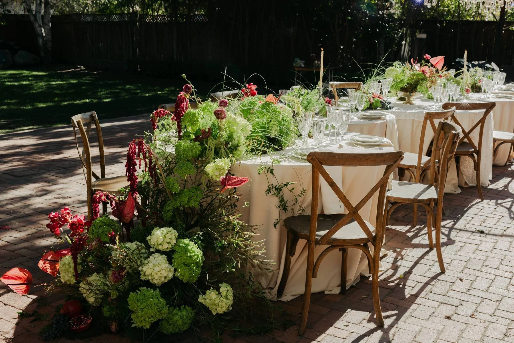 A long outdoor dining table decorated with elegant floral centerpieces and glassware, set up for a celebration or event on a brick patio with sunlit shadows.
