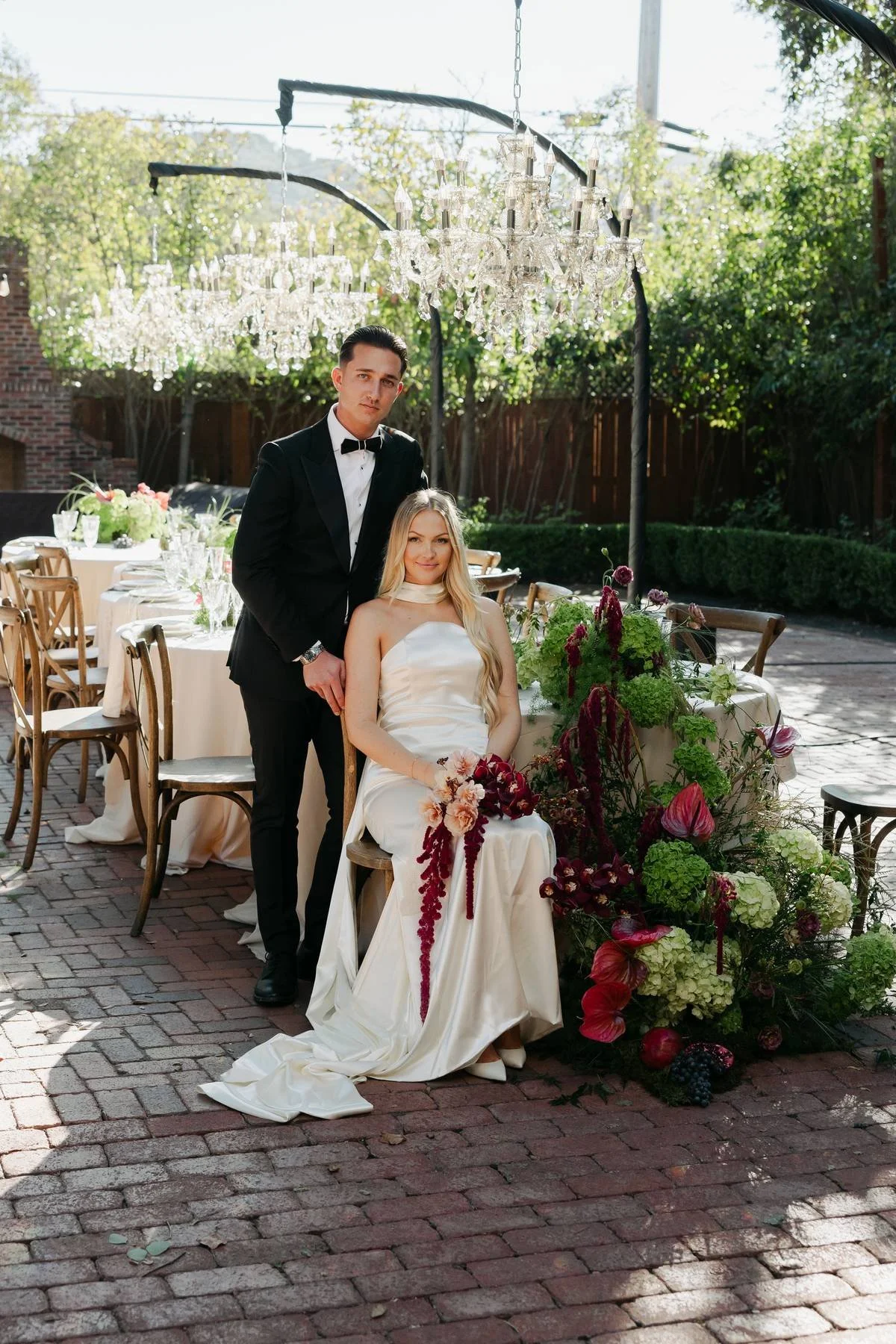 A bride and groom posing outdoors at a wedding reception, with the bride sitting on a wooden chair and the groom standing beside her. They are dressed formally, with chandeliers hanging overhead and decorated tables with floral arrangements in the background.