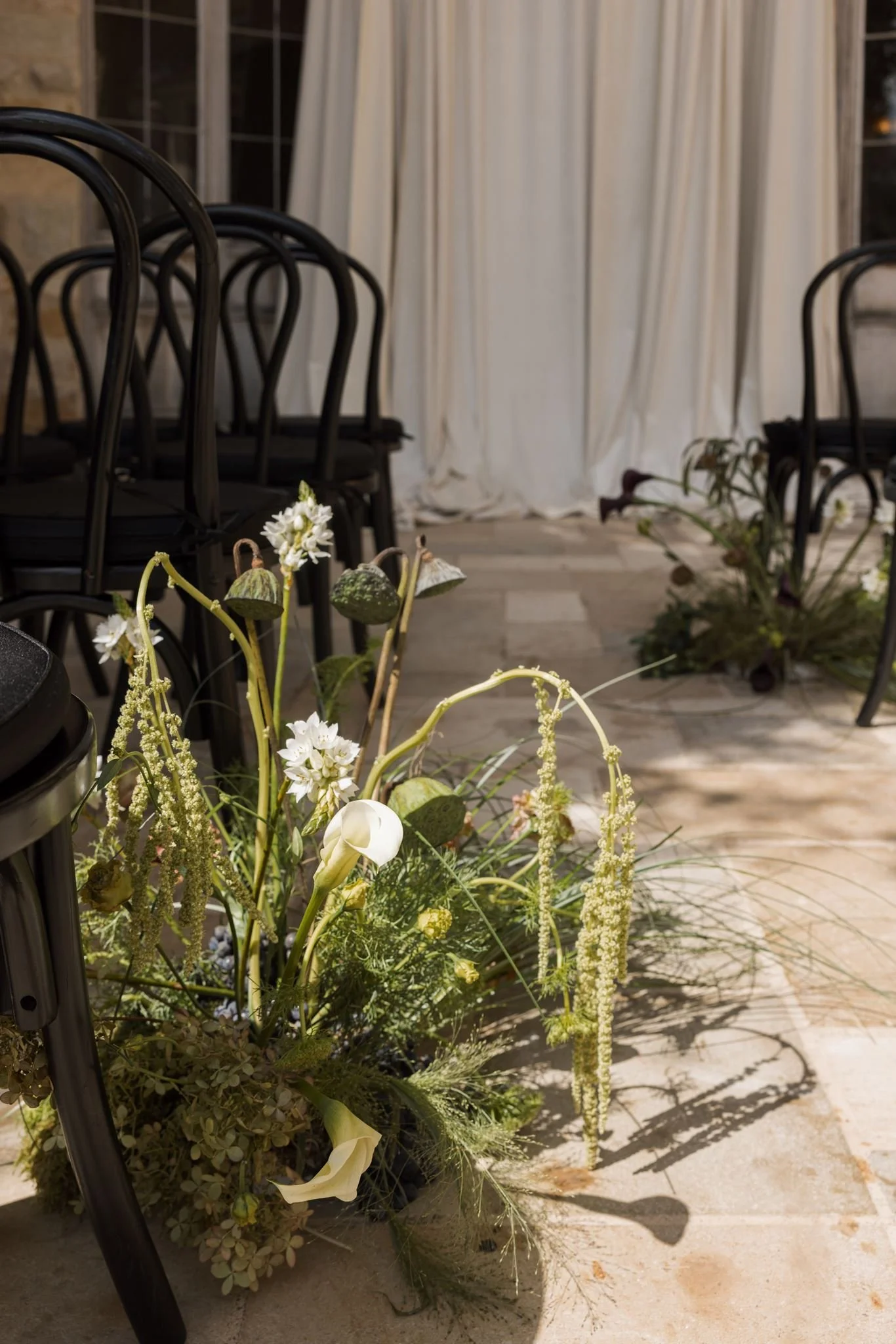 Flower arrangement with white and green flowers and foliage in a black container, placed outdoors near black metal chairs and cream curtains.
