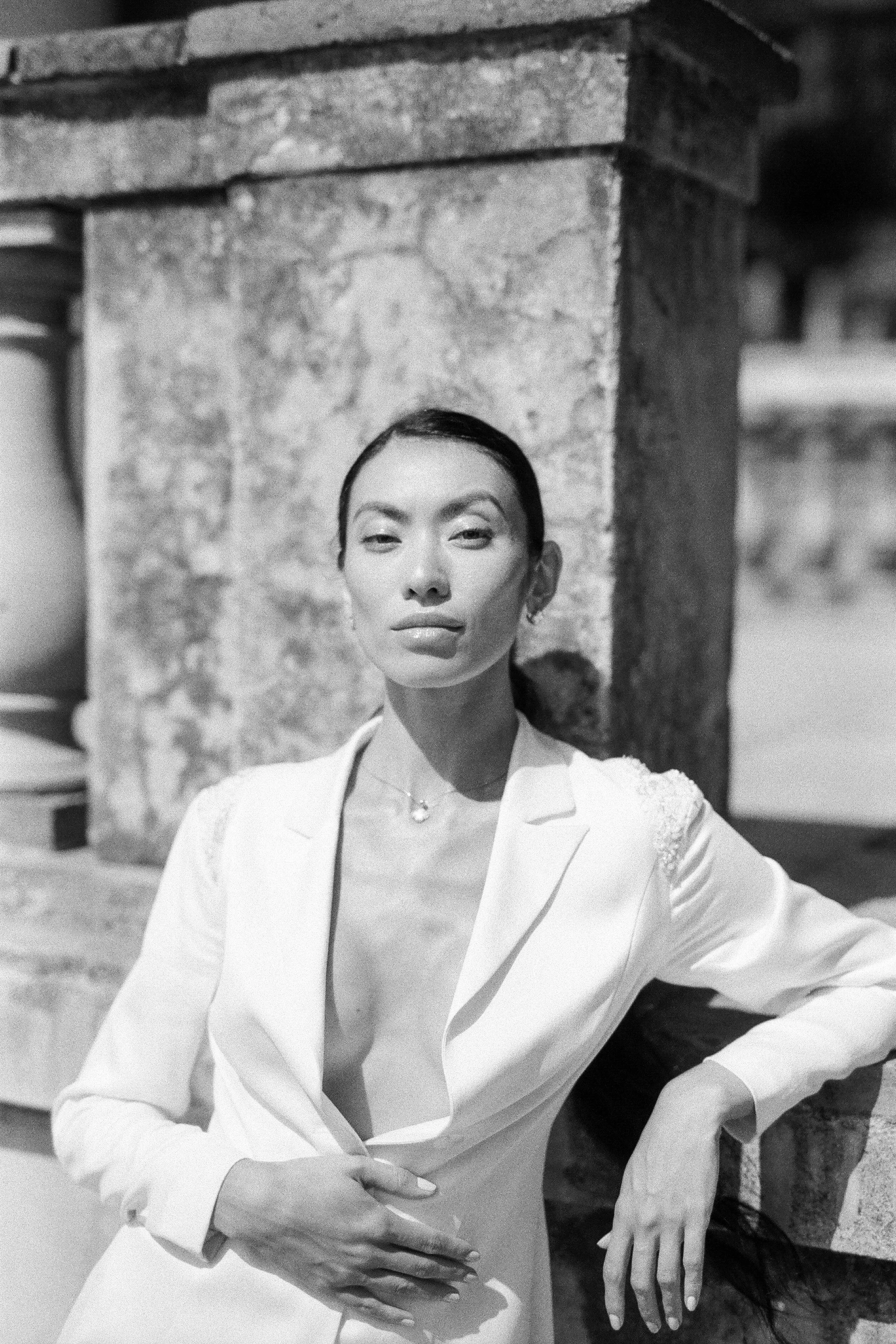 Black and white photograph of a woman wearing a tailored blazer with a low neckline, standing outdoors against a stone wall, with her arms crossed and a confident expression.