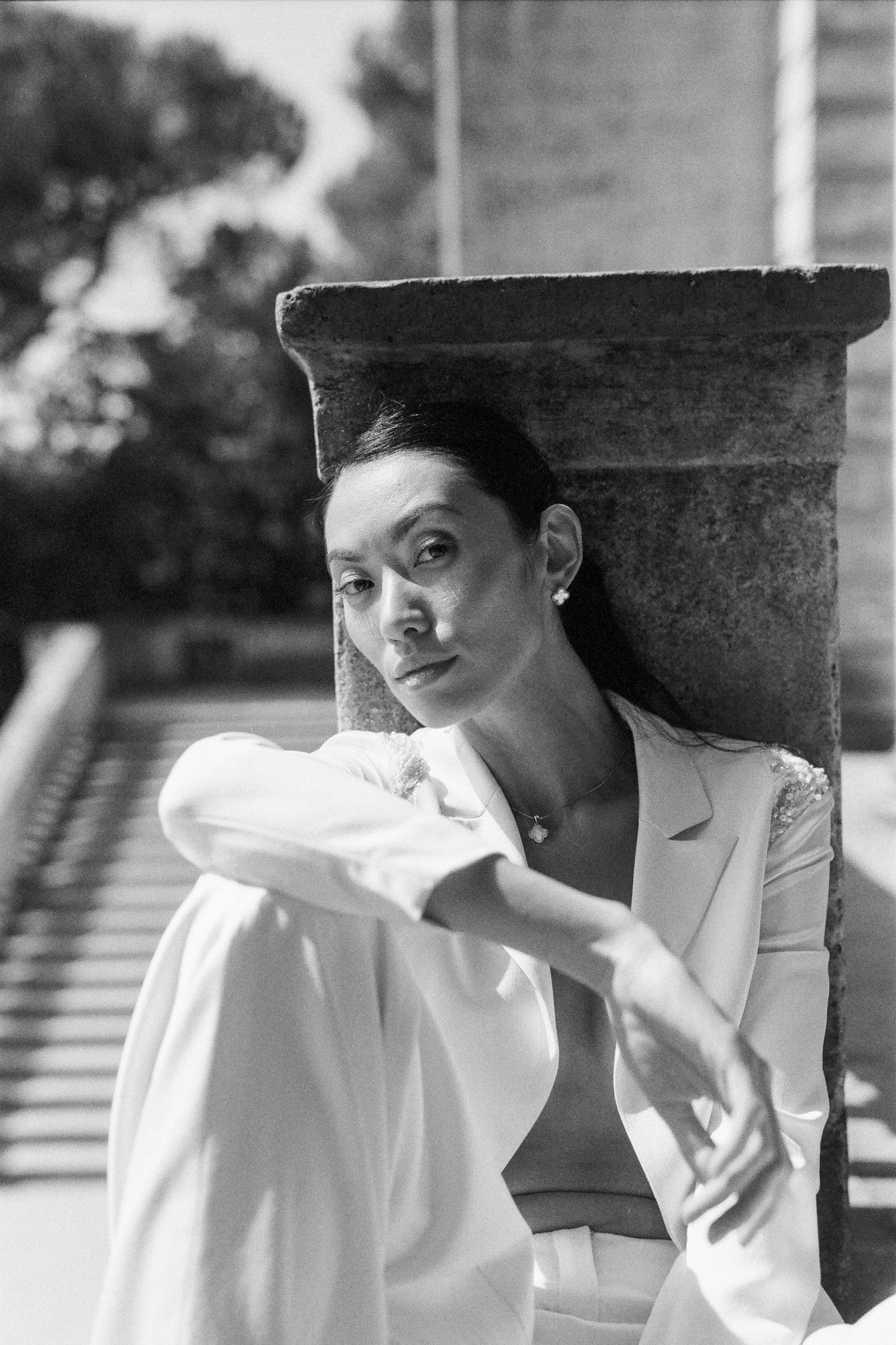 A woman with dark hair tied back, wearing earrings and light-colored clothing, stands near a stucco wall and a fence in an outdoor setting, with trees in the background.