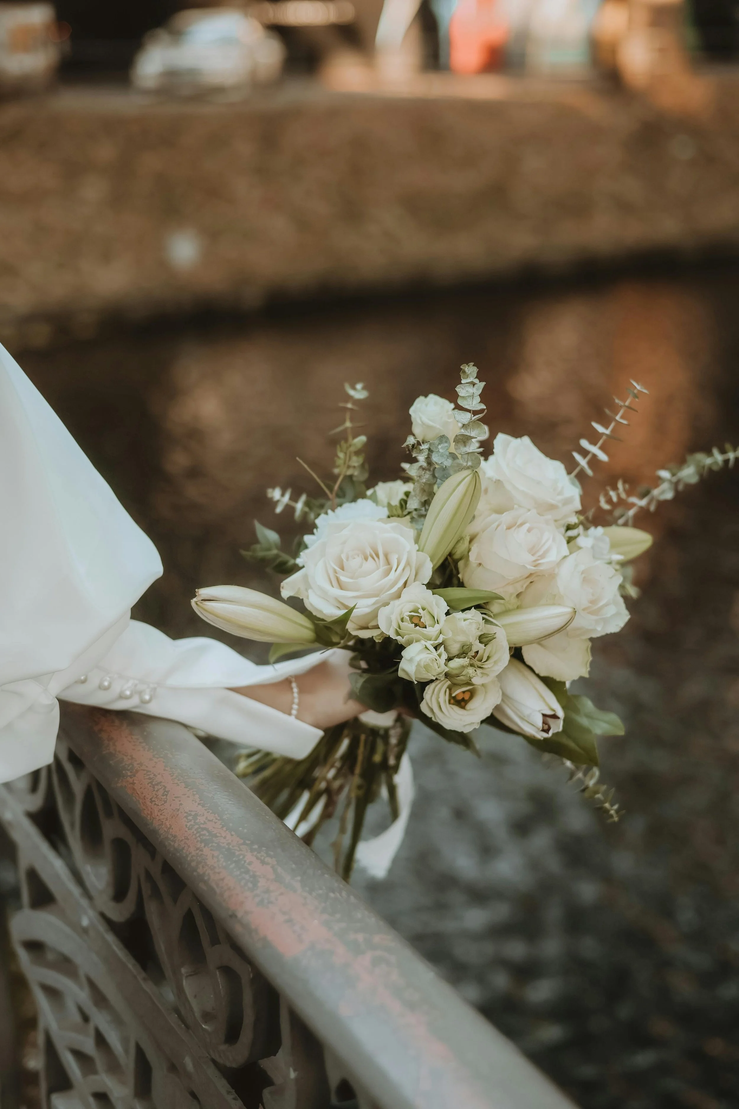 Close-up of a white floral bouquet held by a bride, with a blurred background. The bouquet includes white roses and lilies, with eucalyptus leaves.