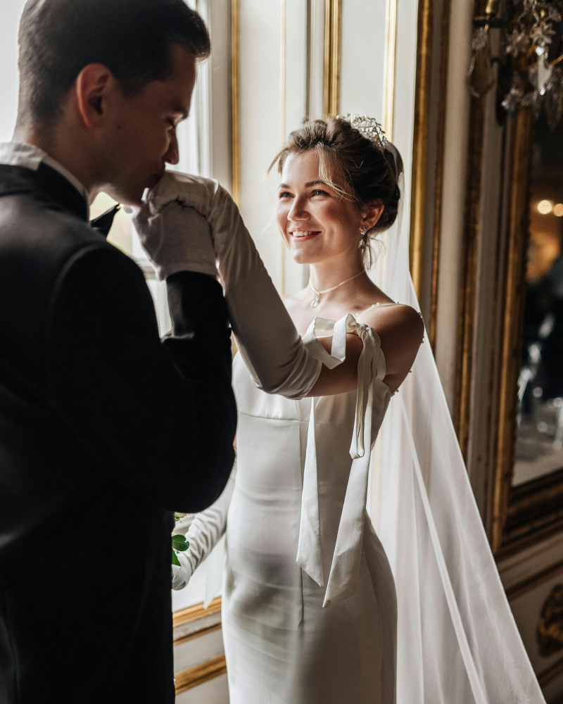A bride with a tiara and veil smiling as a groom looks at her, in an elegant, wood-paneled room with windows and chandeliers.