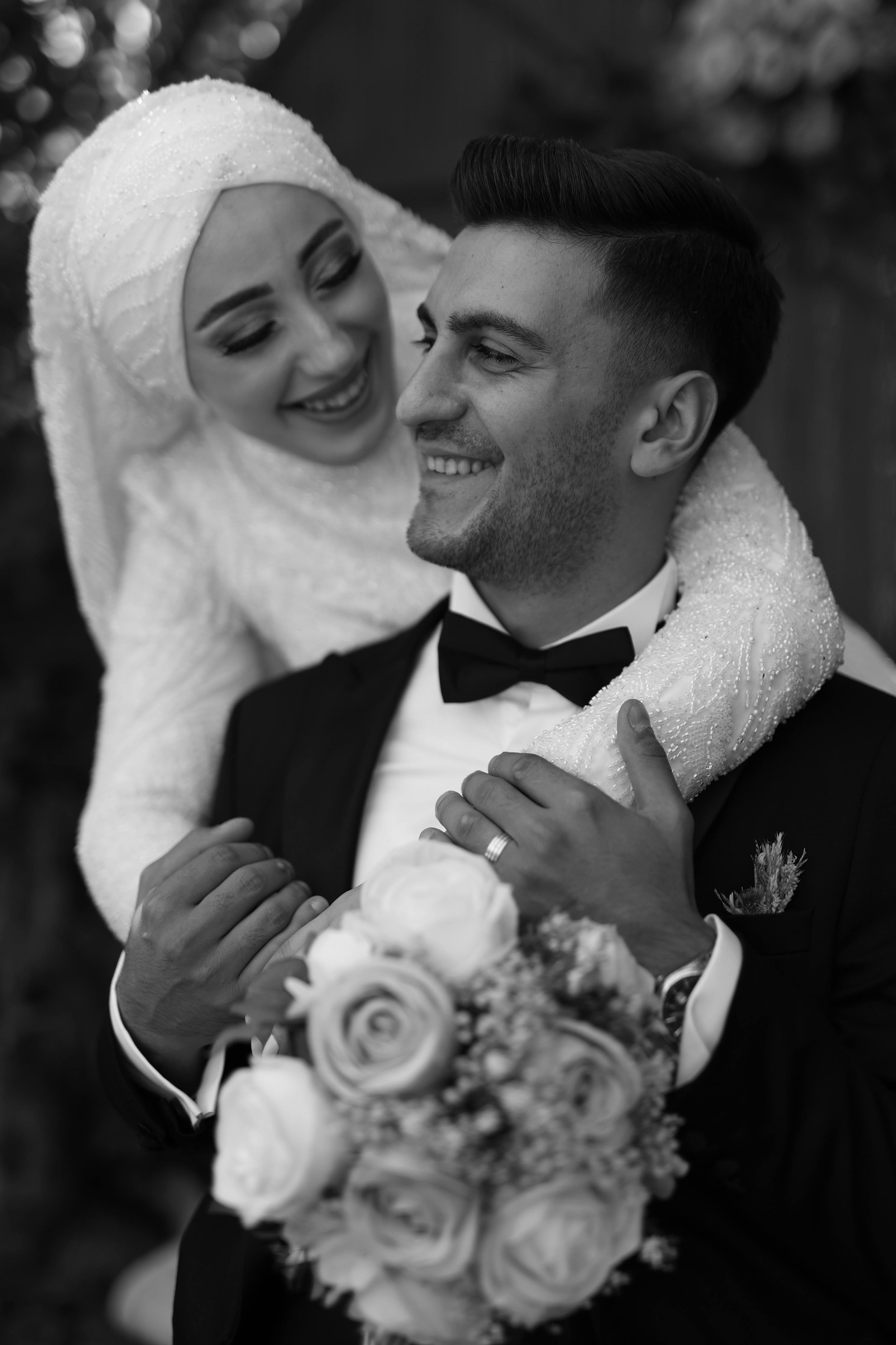 A bride and groom smiling and embracing, with the bride wearing a white headscarf and the groom in a black tuxedo.