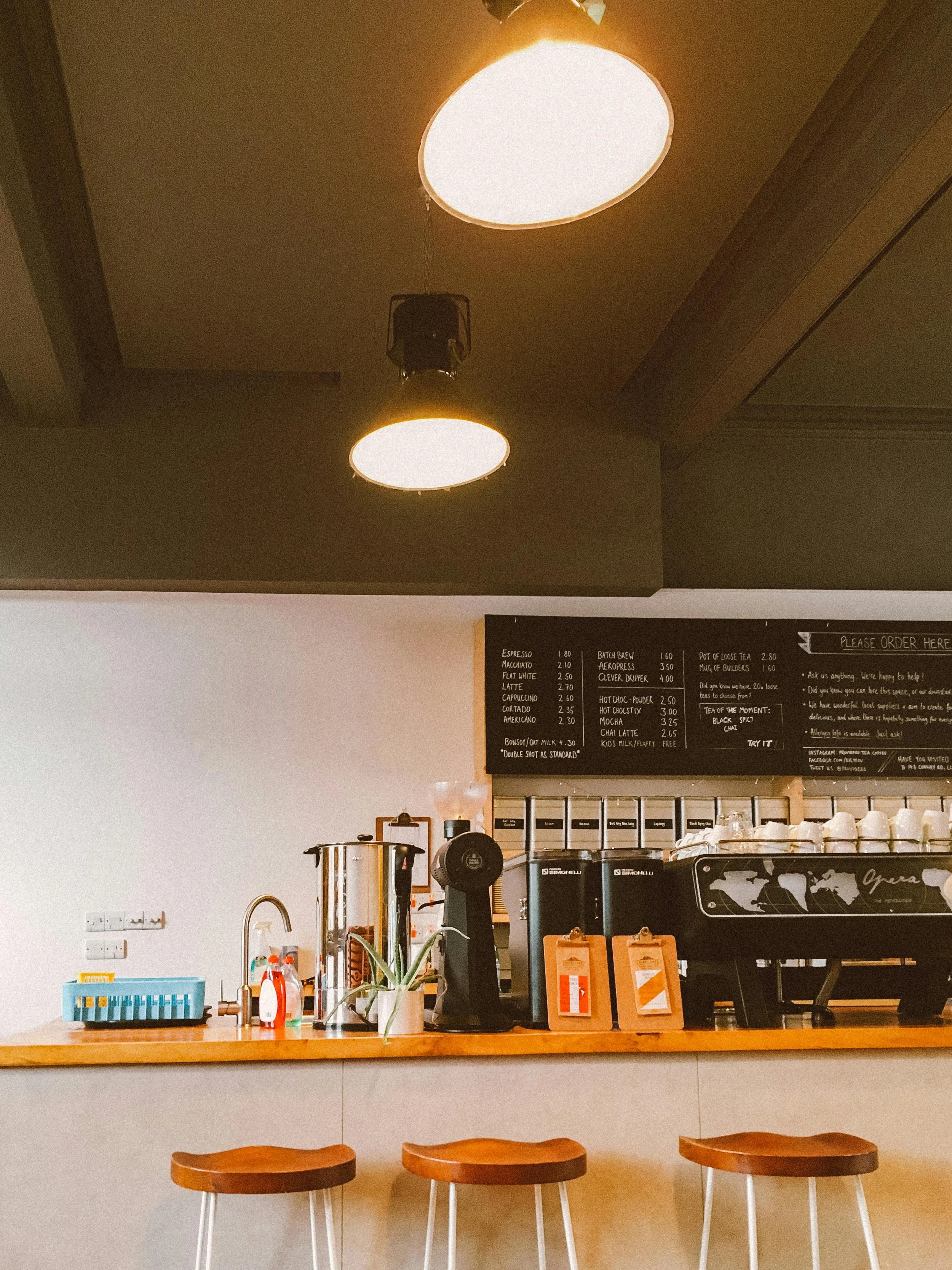 A coffee shop counter with three wooden bar stools below a blackboard menu and oval hanging lights on the ceiling.