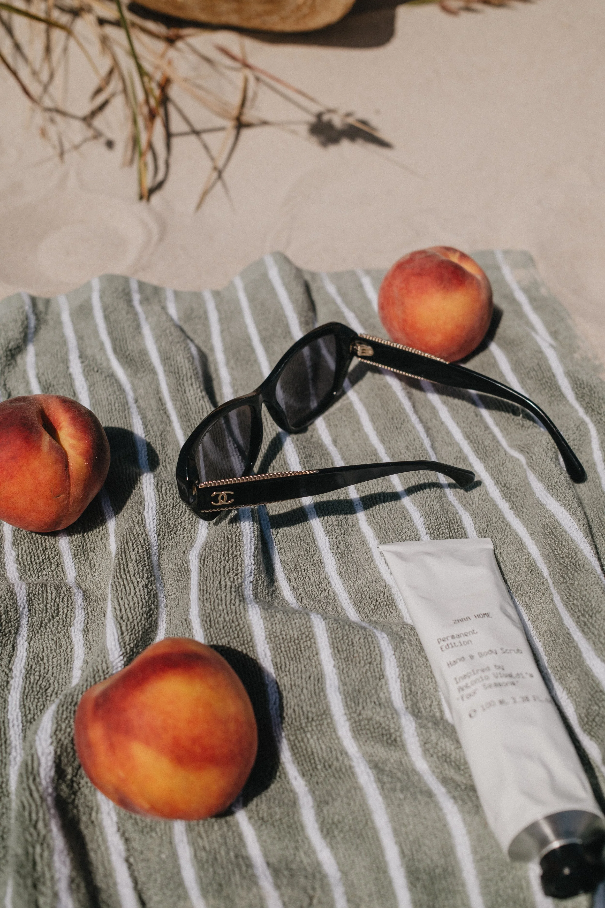 Peach fruits, a pair of black Chanel sunglasses with gold accents, a tube of ZARA Home skincare product, and a striped towel on a surface with some dried plants in the background.