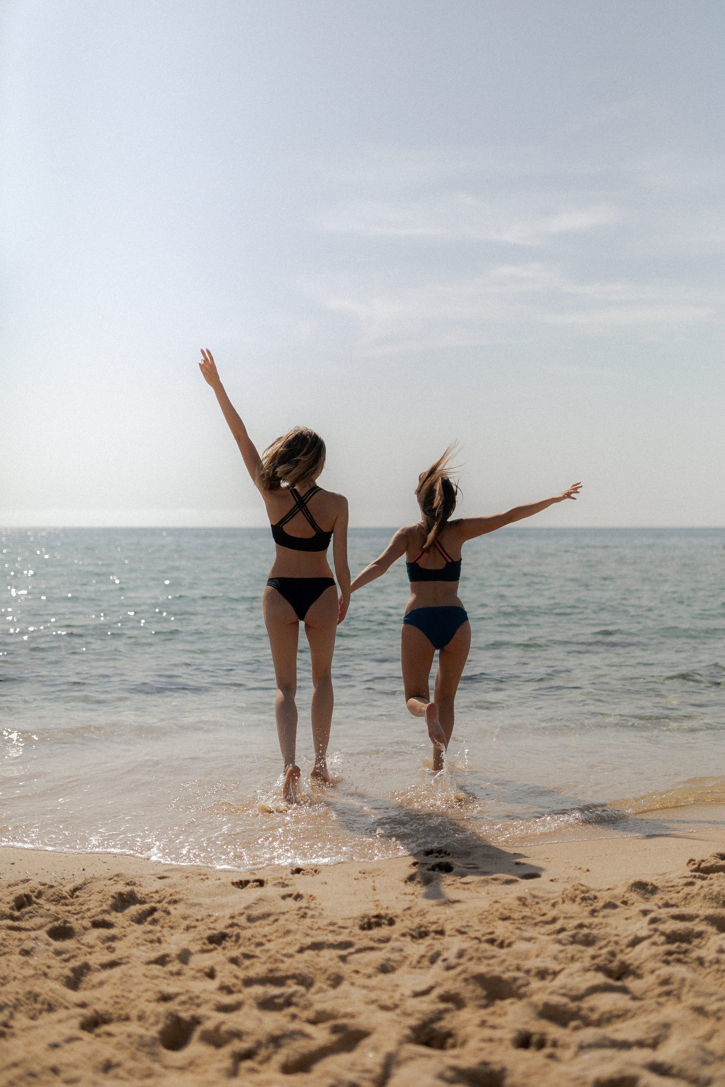 Two women in bikinis holding hands and running into the ocean at the beach with the sky and water in the background.