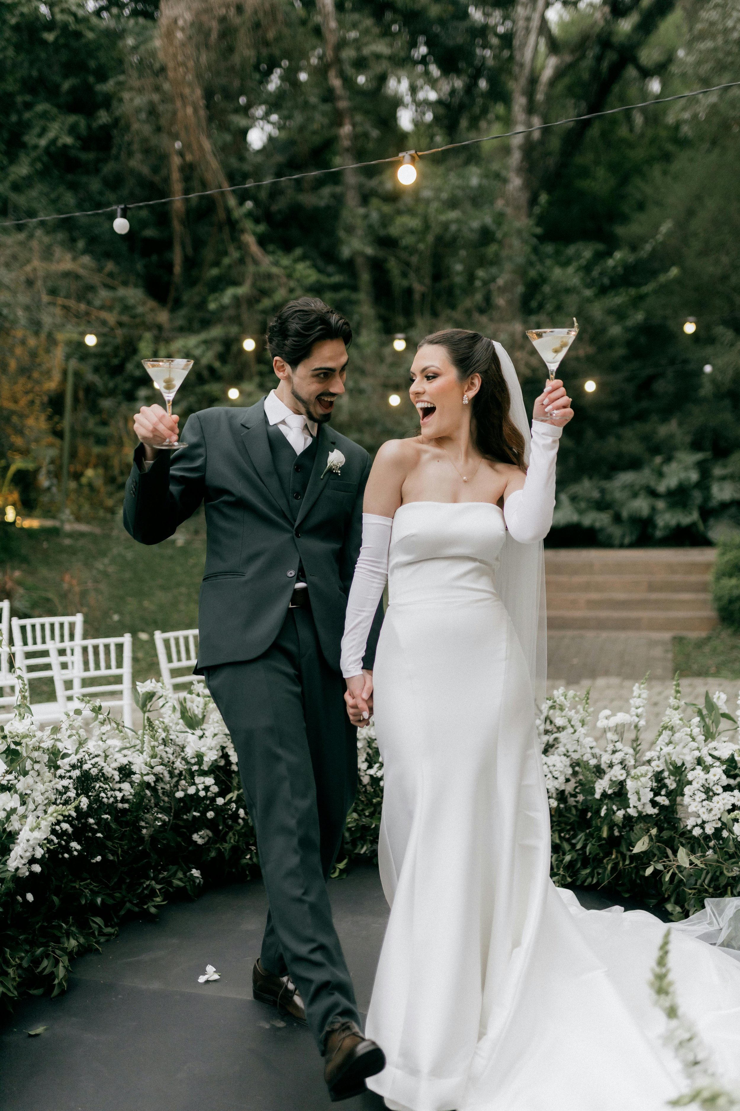 A bride and groom holding hands and toasting with martini glasses at their outdoor wedding ceremony with fairy lights overhead and white flowers along the aisle.