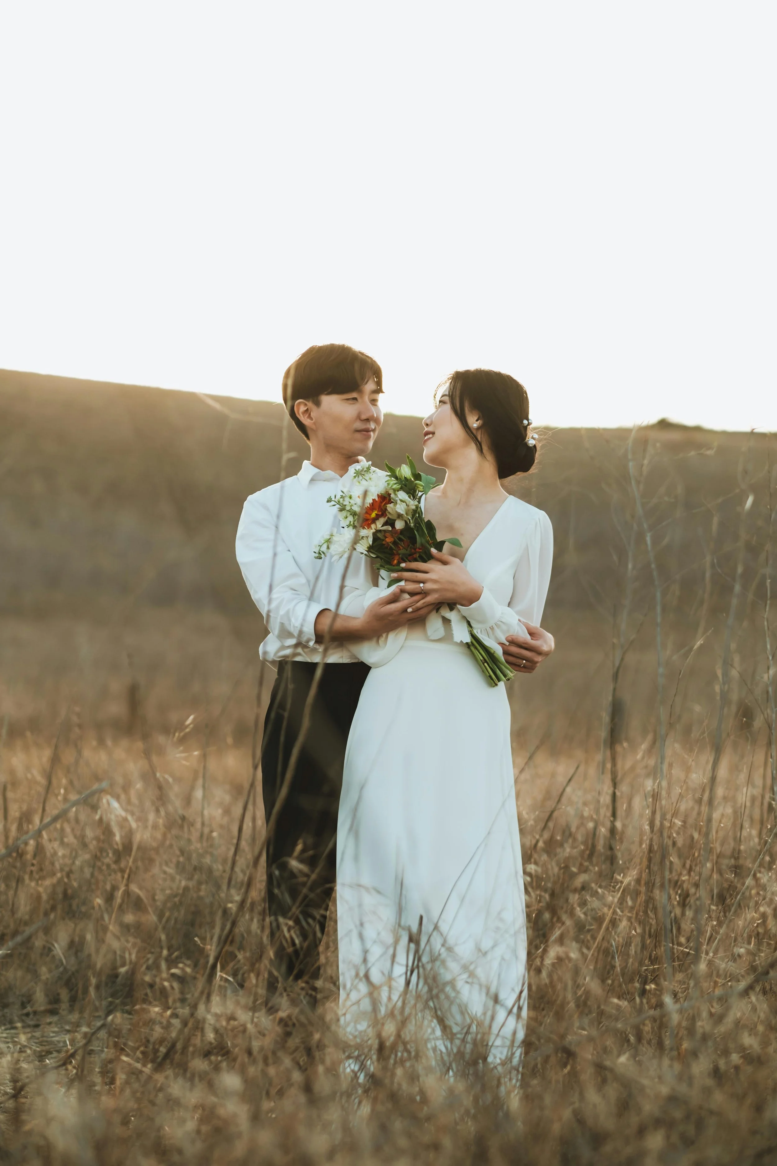 A man and woman stand close together in a field of tall, dry grass during sunset. The man is dressed in a white shirt, while the woman wears a white dress and holds a bouquet of flowers. They look into each other's eyes with a romantic expression.