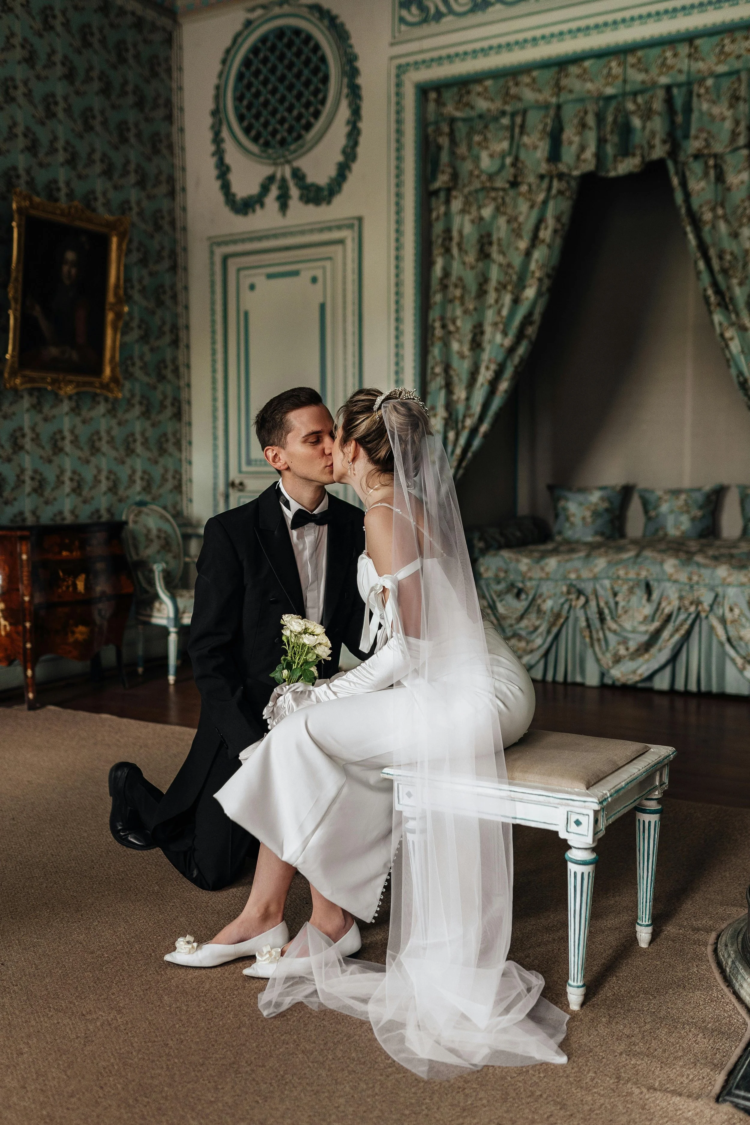 A bride and groom sharing a kiss indoors in an elegant room. The groom, dressed in a black tuxedo with a bow tie, is kneeling on one knee, holding a bouquet of white roses. The bride, wearing a white wedding dress and veil, is seated on a cream-colored bench. The room has ornate wallpaper, decorative curtains, and framed artwork.