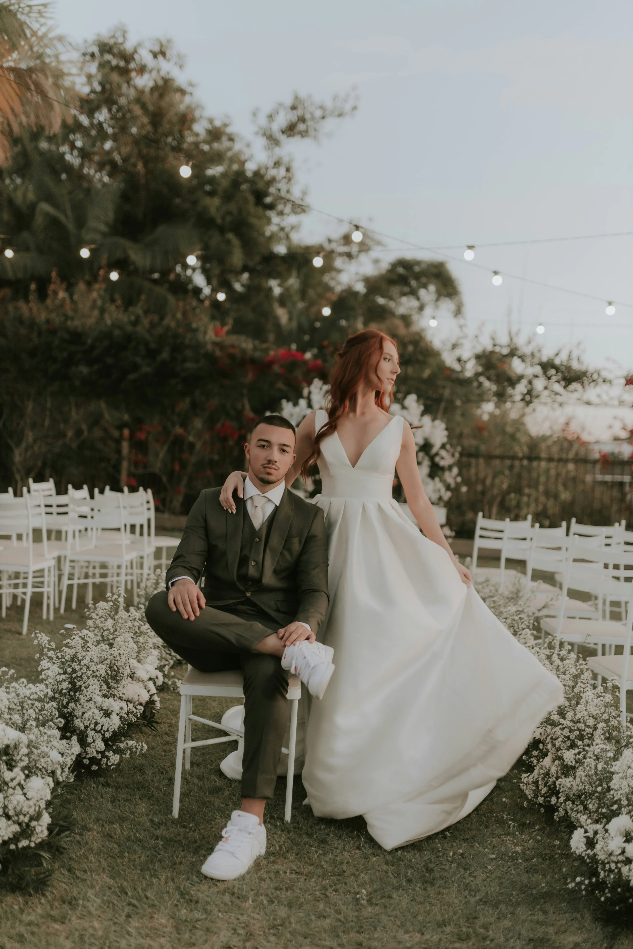 A man in a suit seated on a white chair and a woman in a wedding gown standing behind him outdoors during evening, surrounded by white flowers and chairs, with string lights hanging in the background.
