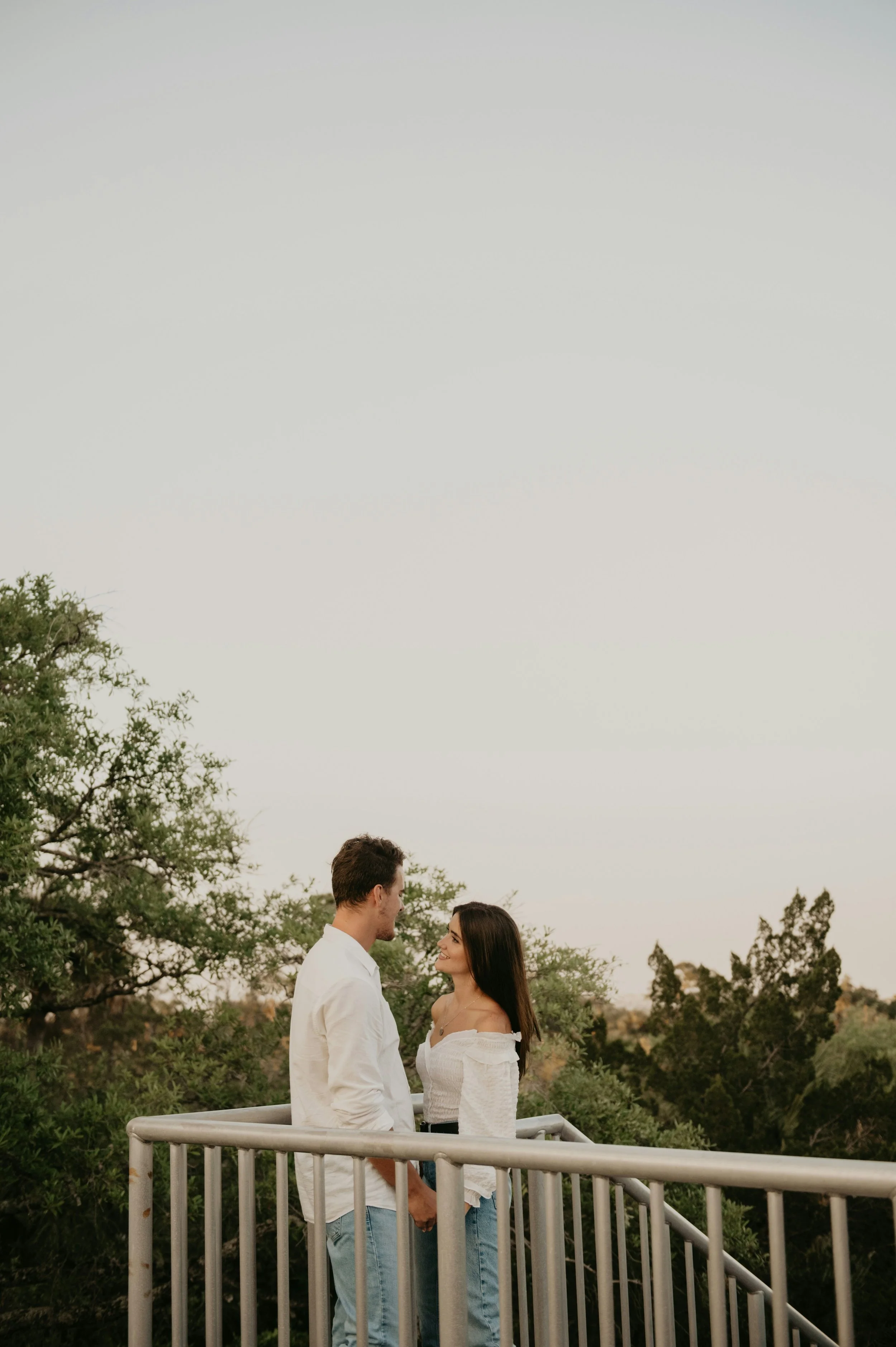 A couple stands on a balcony holding hands, gazing into each other's eyes, surrounded by trees and an overcast sky.
