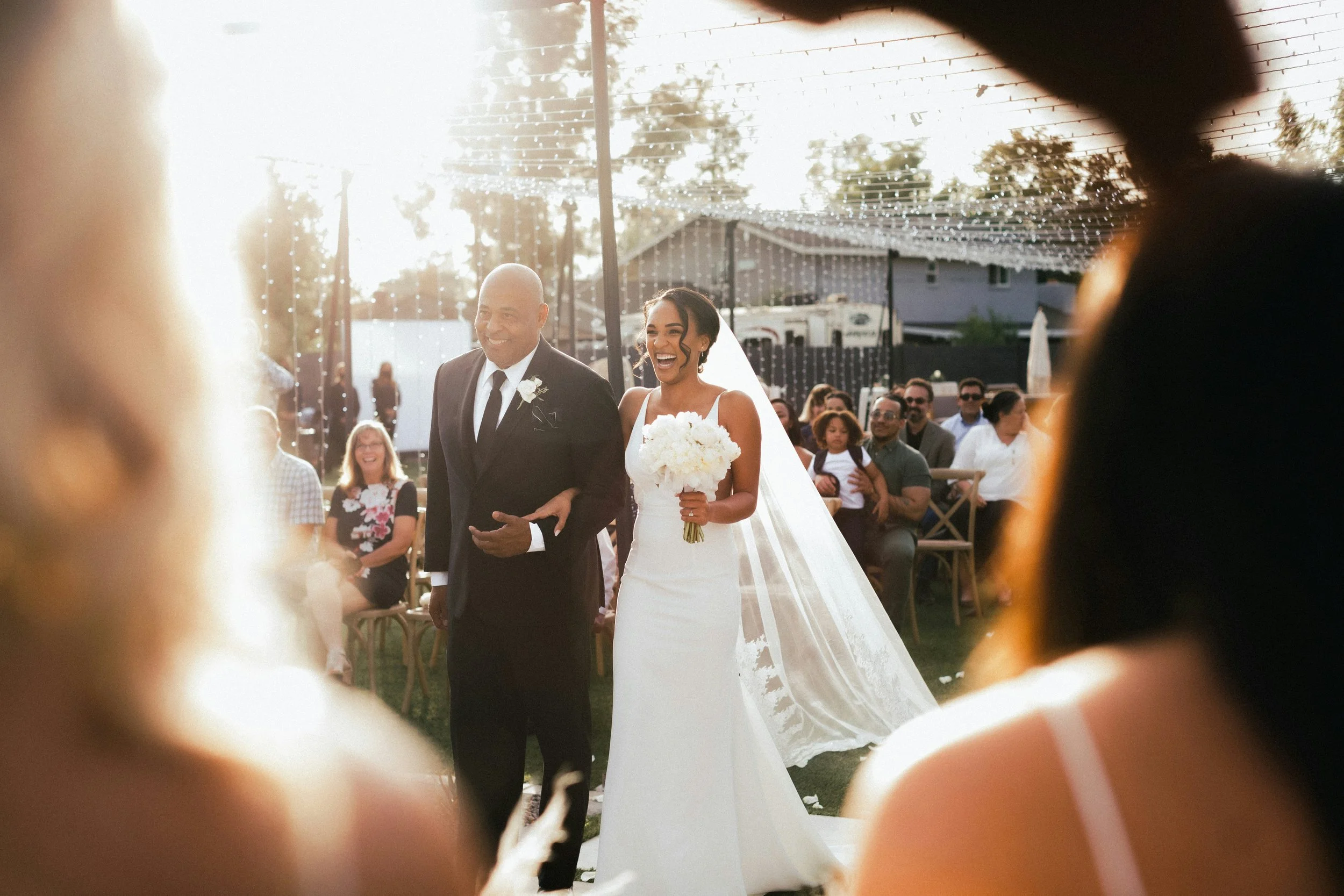 A wedding ceremony with a bride in a white dress and bouquet walking down the aisle, accompanied by a man in a dark suit, with guests seated outdoors under string lights at sunset.