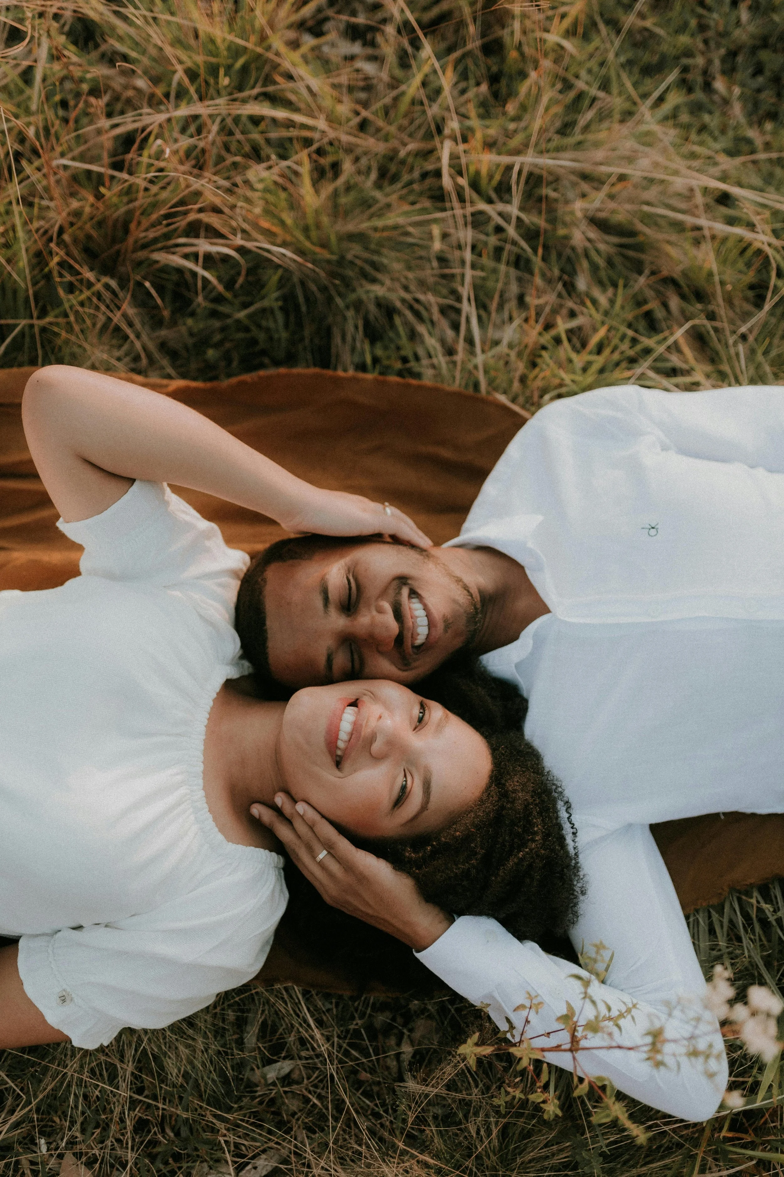 Two smiling people, one man with light skin and one woman with darker skin, lying on the ground amidst grass and small plants. They have relaxed expressions and are wearing white tops.