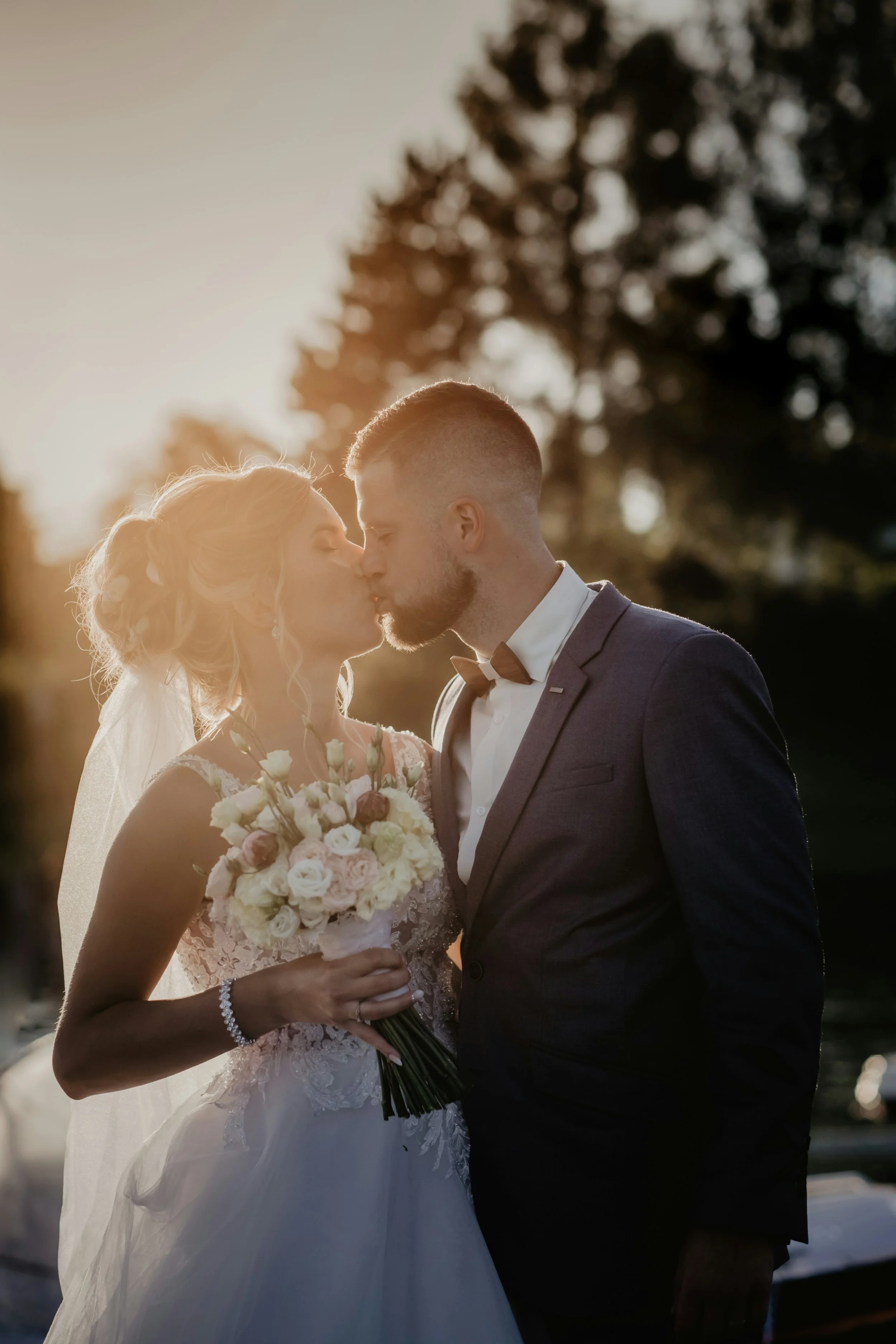 A bride and groom sharing a kiss outdoors during sunset, with the bride holding a bouquet of white and pink roses, the bride wearing a lace wedding gown, and the groom in a dark suit and bow tie.