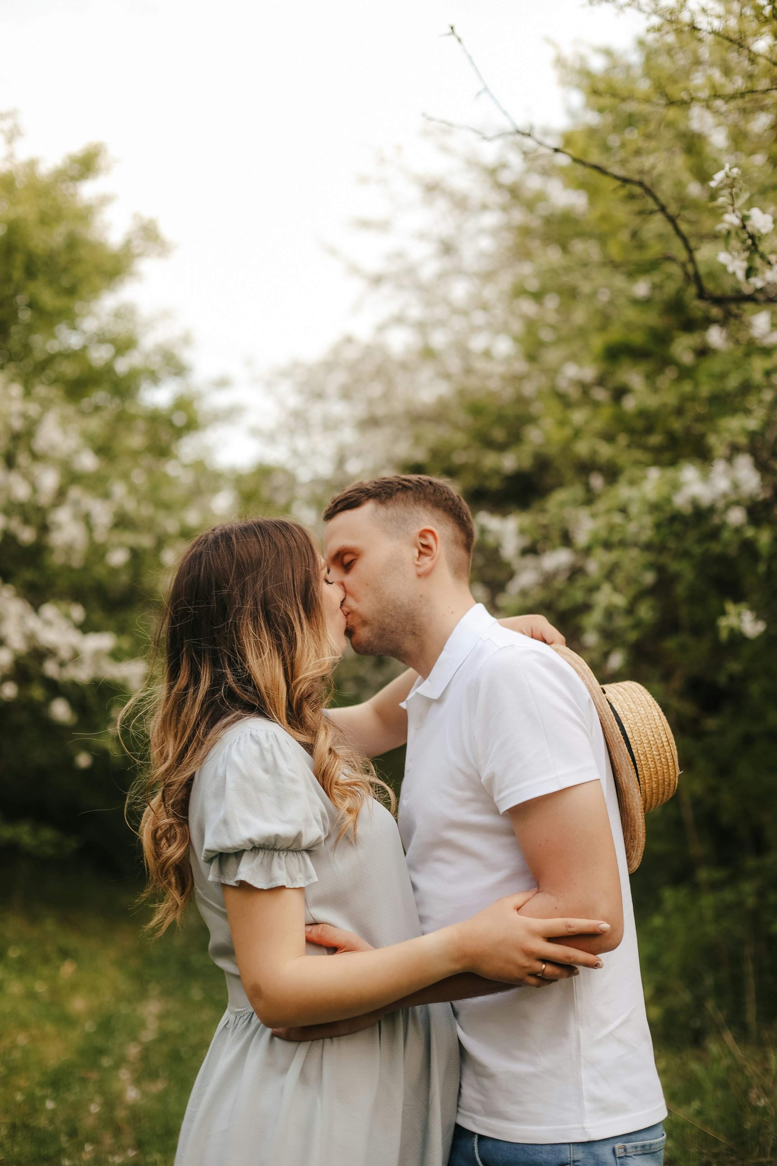 A young couple is kissing outdoors during spring, with trees in bloom behind them.