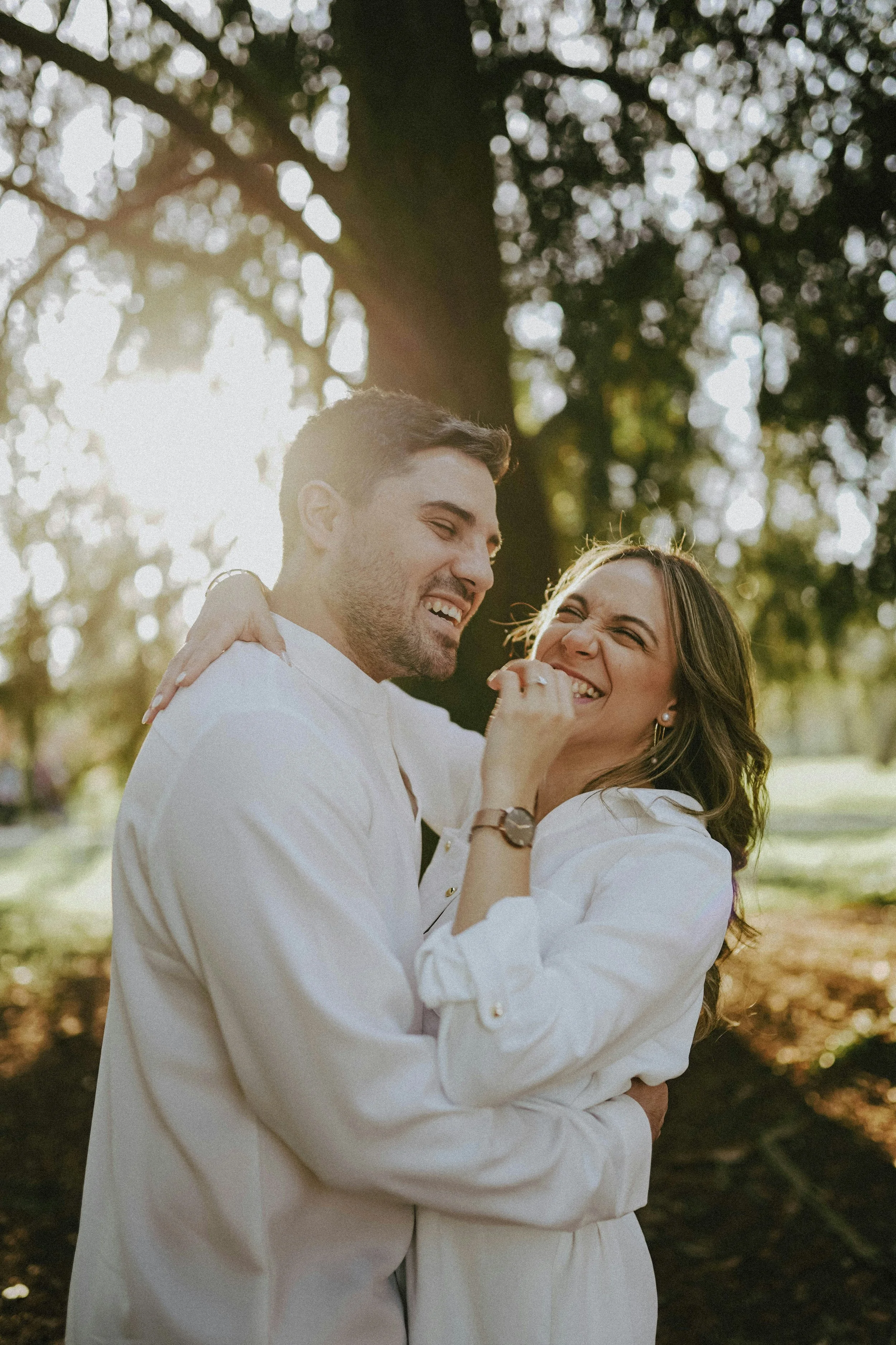 A joyful couple embracing outdoors in a park during daytime, smiling and laughing with sunlight filtering through trees.