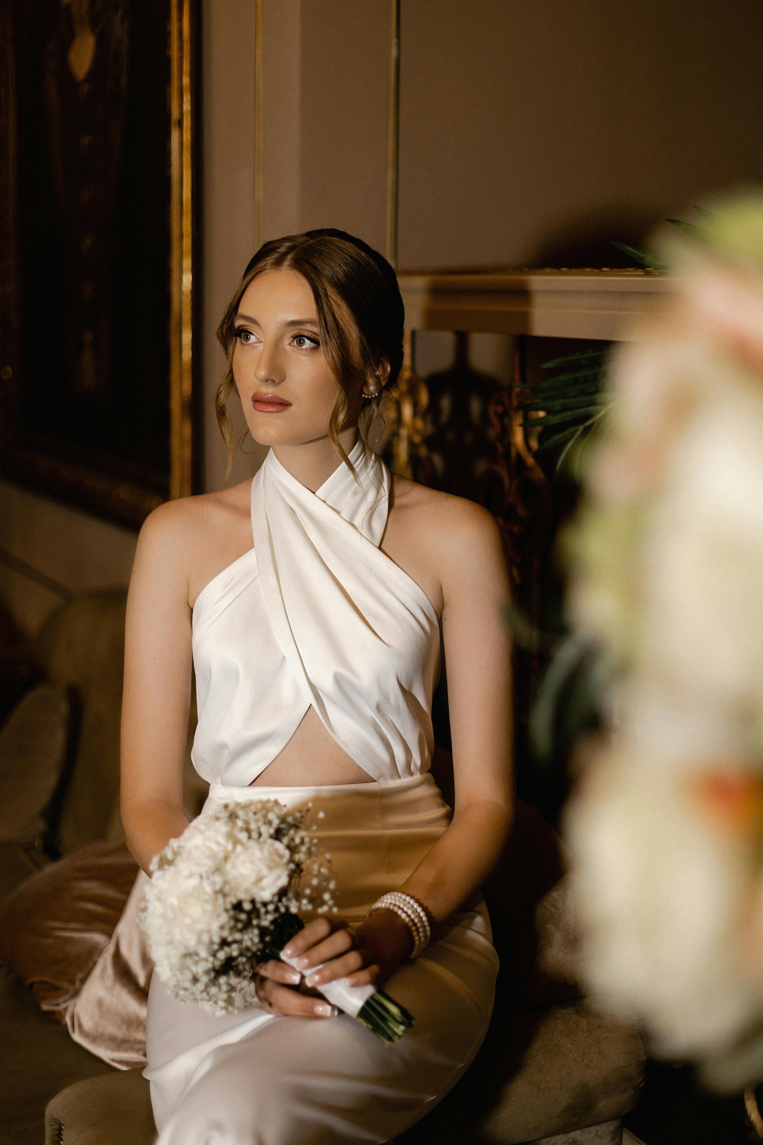 A young woman dressed in elegant attire, sitting indoors holding a bouquet of white flowers, with soft lighting and ornate decor in the background.