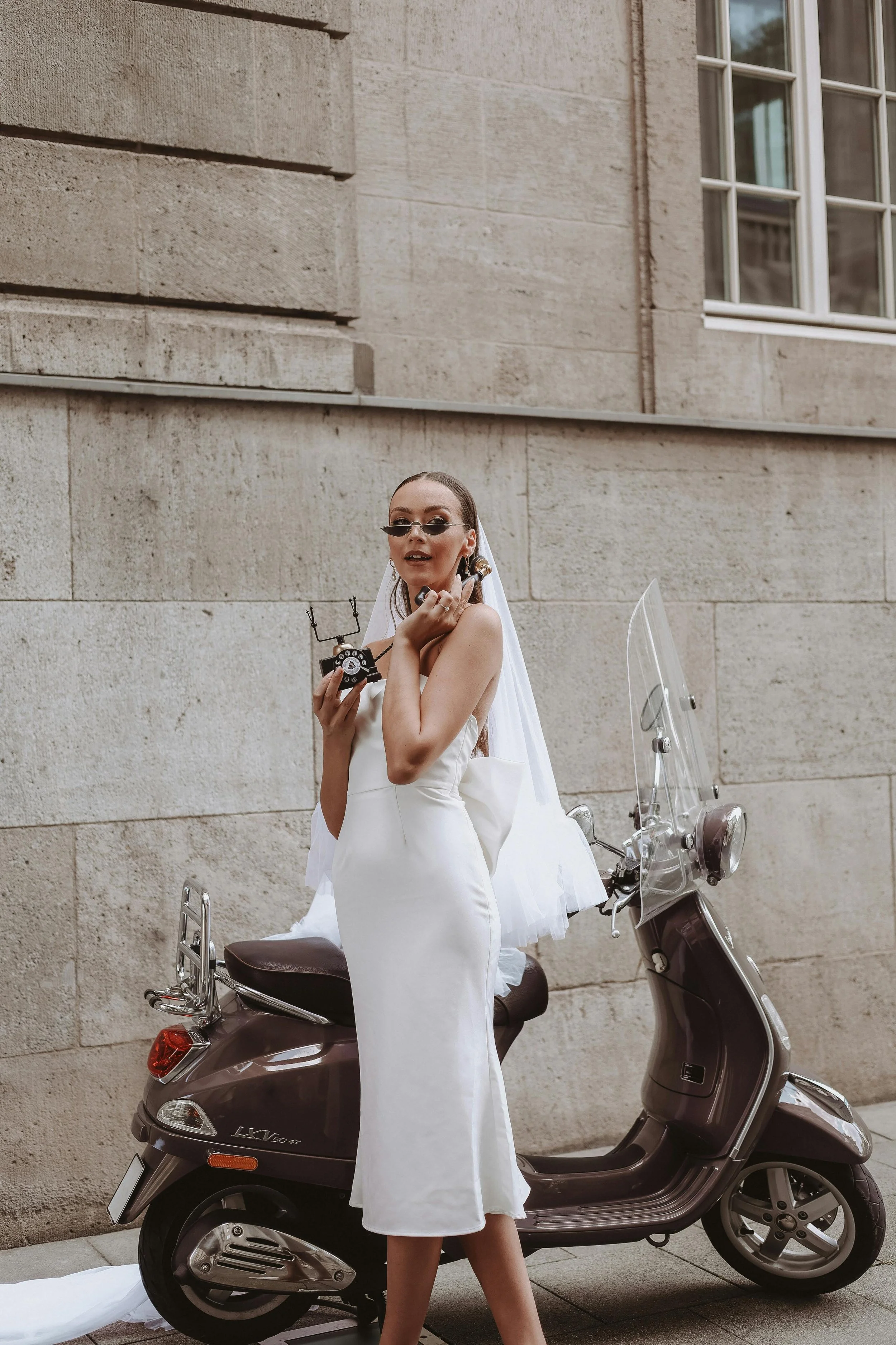 A woman in a white dress with sunglasses, holding a vintage camera, standing in front of a brown scooter parked against a beige stone wall with a window.