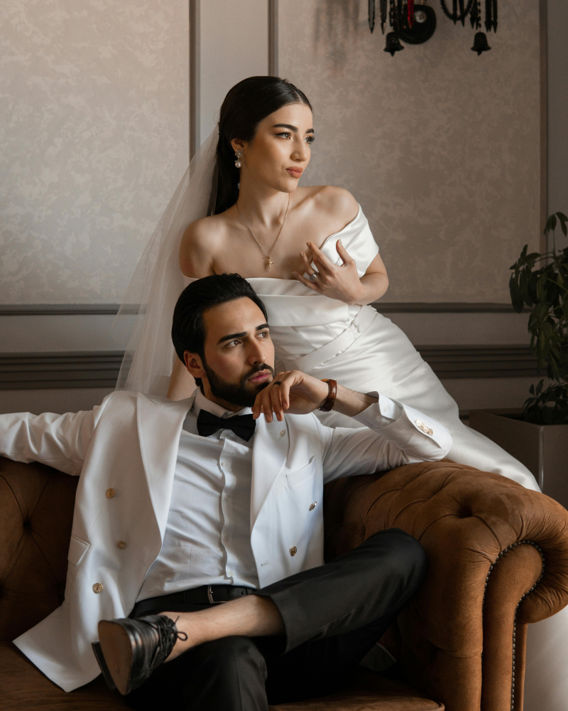 A bride and groom in formal wedding attire posing indoors. The bride is standing behind the groom, wearing an off-the-shoulder white dress and veil, with jewelry. The groom, seated on a vintage brown leather sofa, is wearing a white tuxedo jacket wit