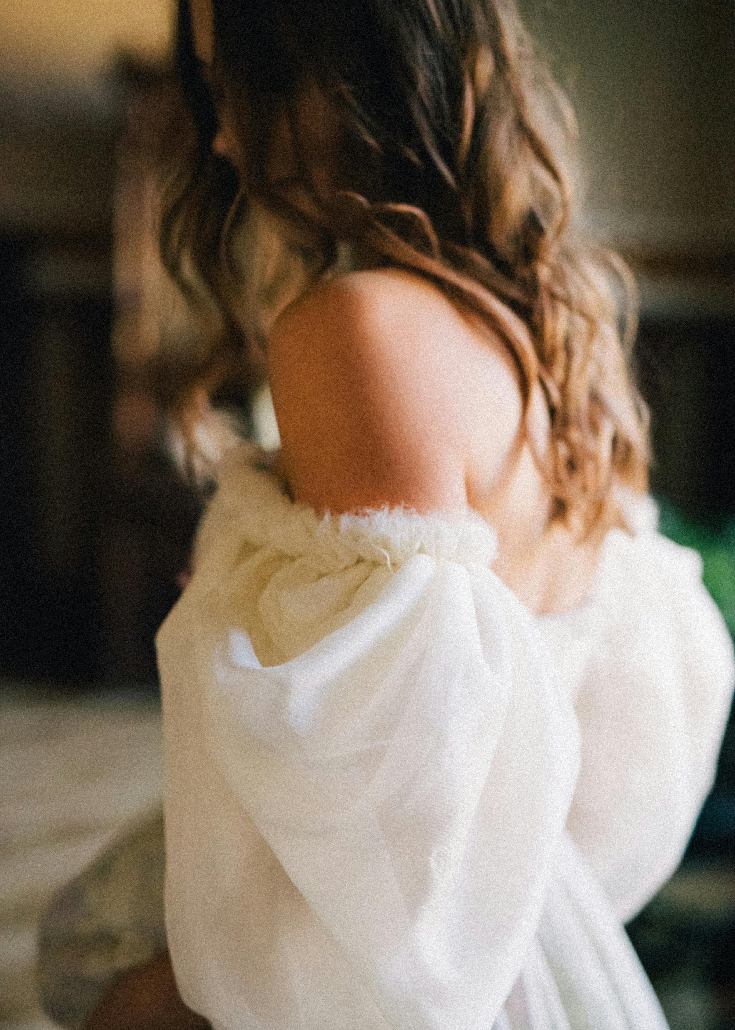 Close-up of a woman's shoulder with brown, curly hair, wearing a white, fluffy dress with a bow detail.