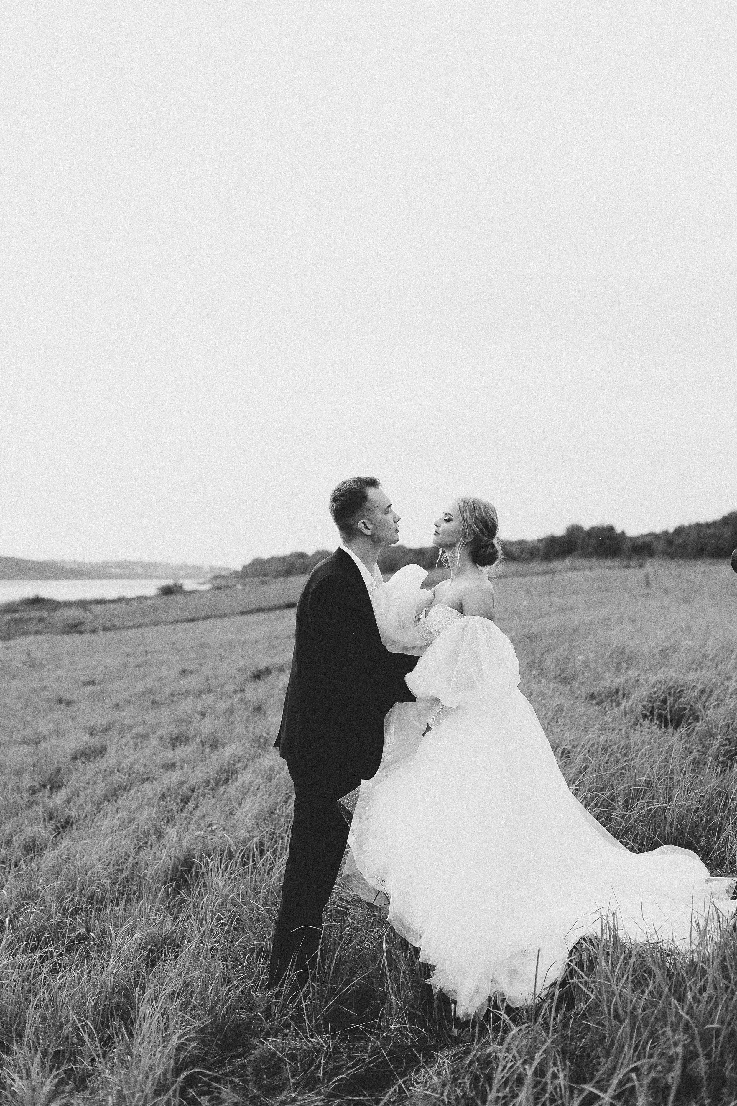 A black-and-white photo of a bride and groom standing in a grassy field near a body of water, holding hands and gazing into each other's eyes during a wedding photo shoot.