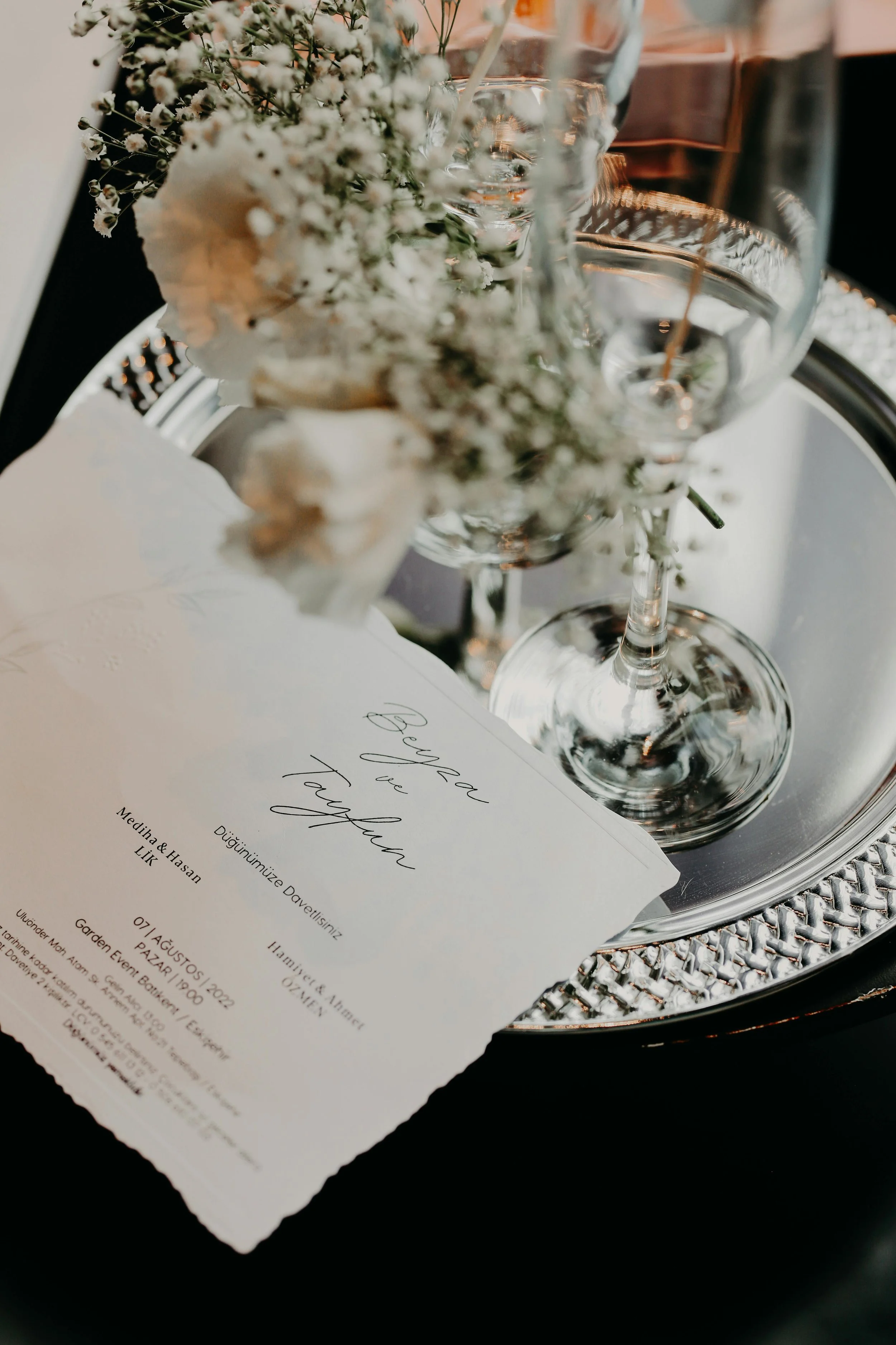 Close-up of a wedding invitation card placed on a silver tray surrounded by wine glasses and a floral arrangement with white flowers and greenery.