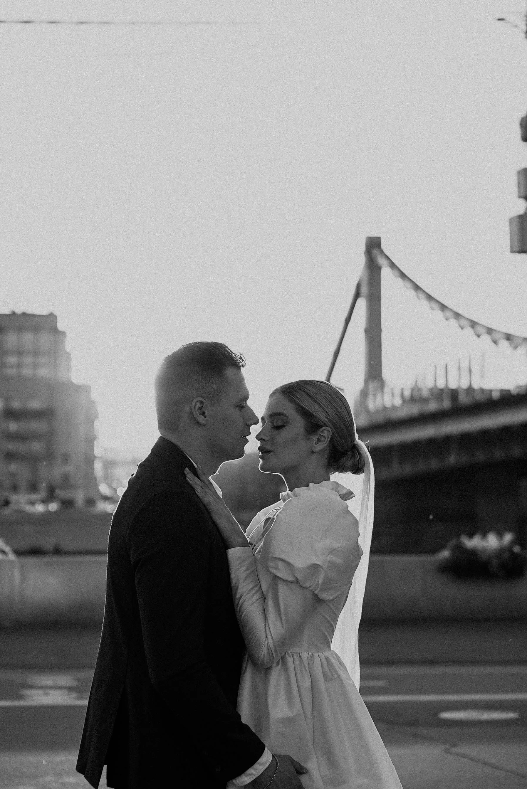 A black and white photo of a couple standing close together outdoors, with a bridge in the background.