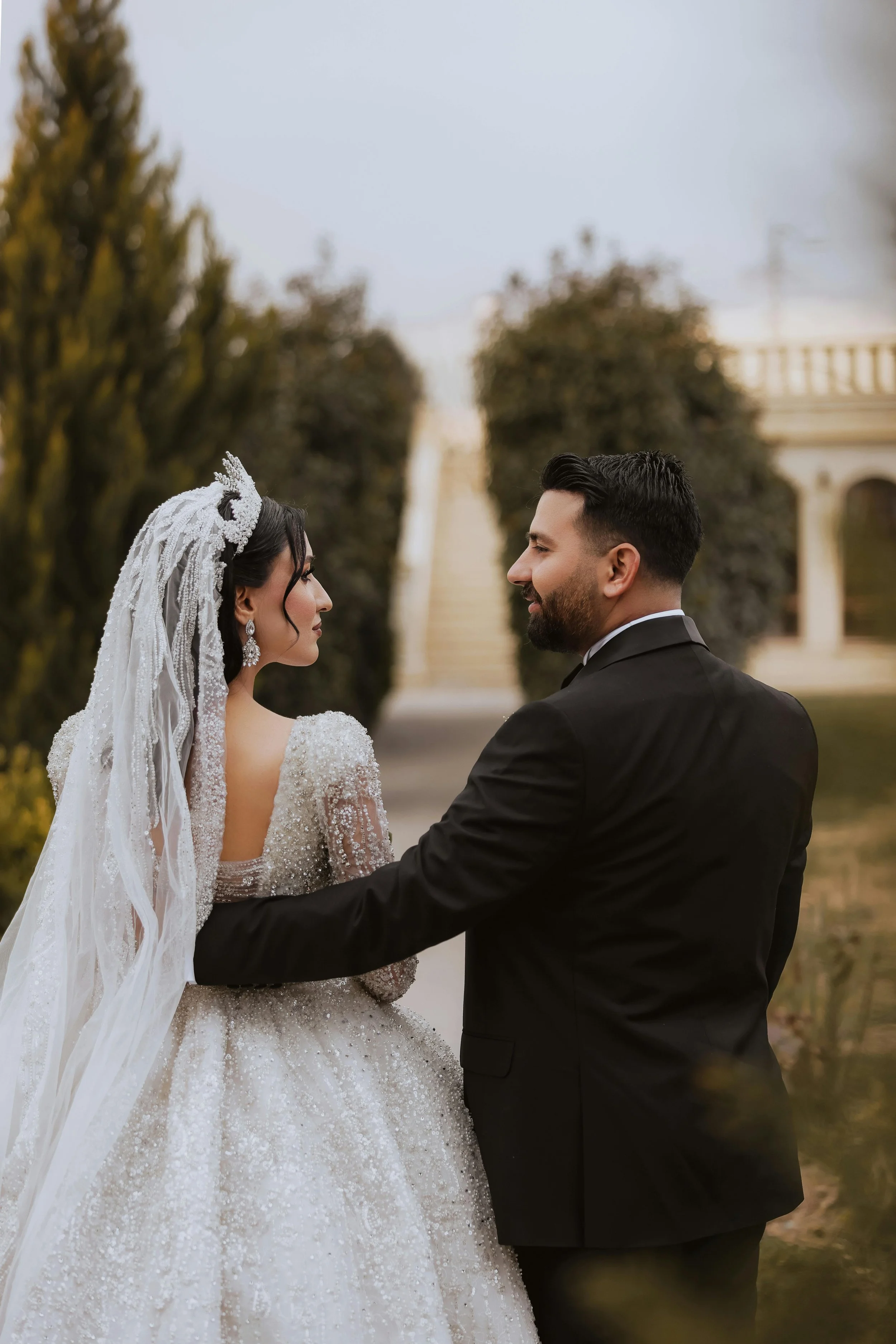 A bride and groom stand facing each other outdoors, holding hands, with a blurred background of trees and a building.