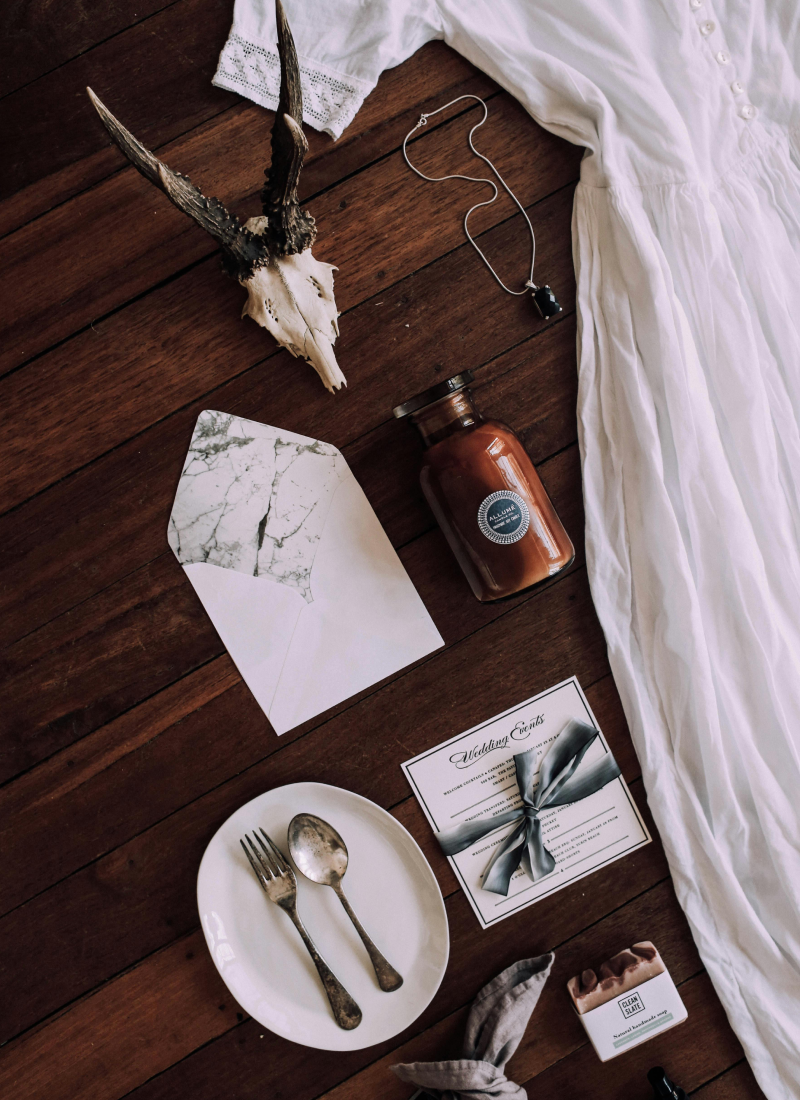 Flat lay of various items on a wooden surface, including a deer skull with antlers, a white dress, a necklace, a brown bottle, an envelope, a wedding invitation with a gray ribbon, a white plate with vintage fork and spoon, a small box of soap, and a cloth napkin.