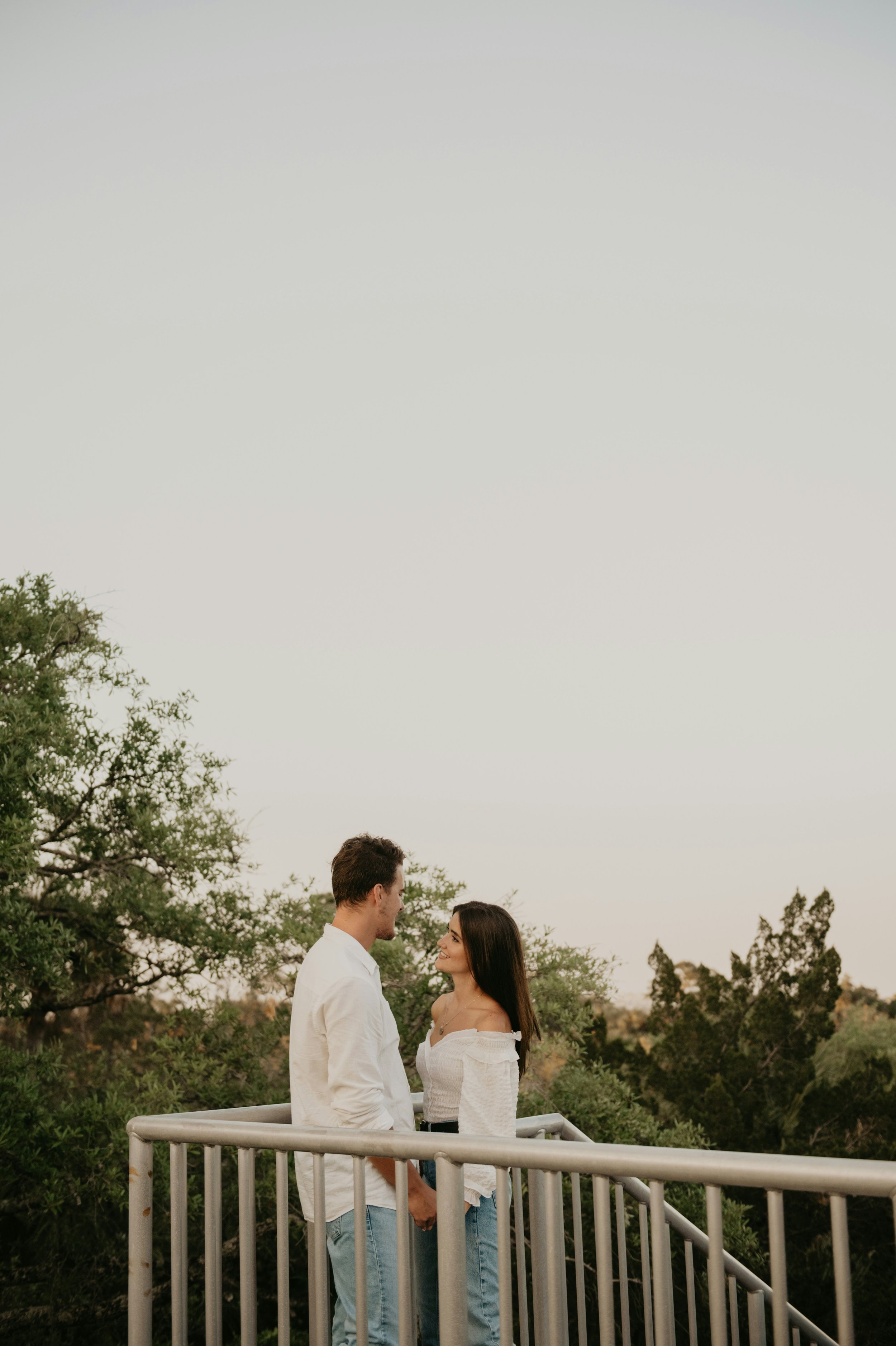 A couple holding hands, standing close to each other on a balcony outdoors, facing each other with happy expressions, surrounded by trees and greenery during sunset.