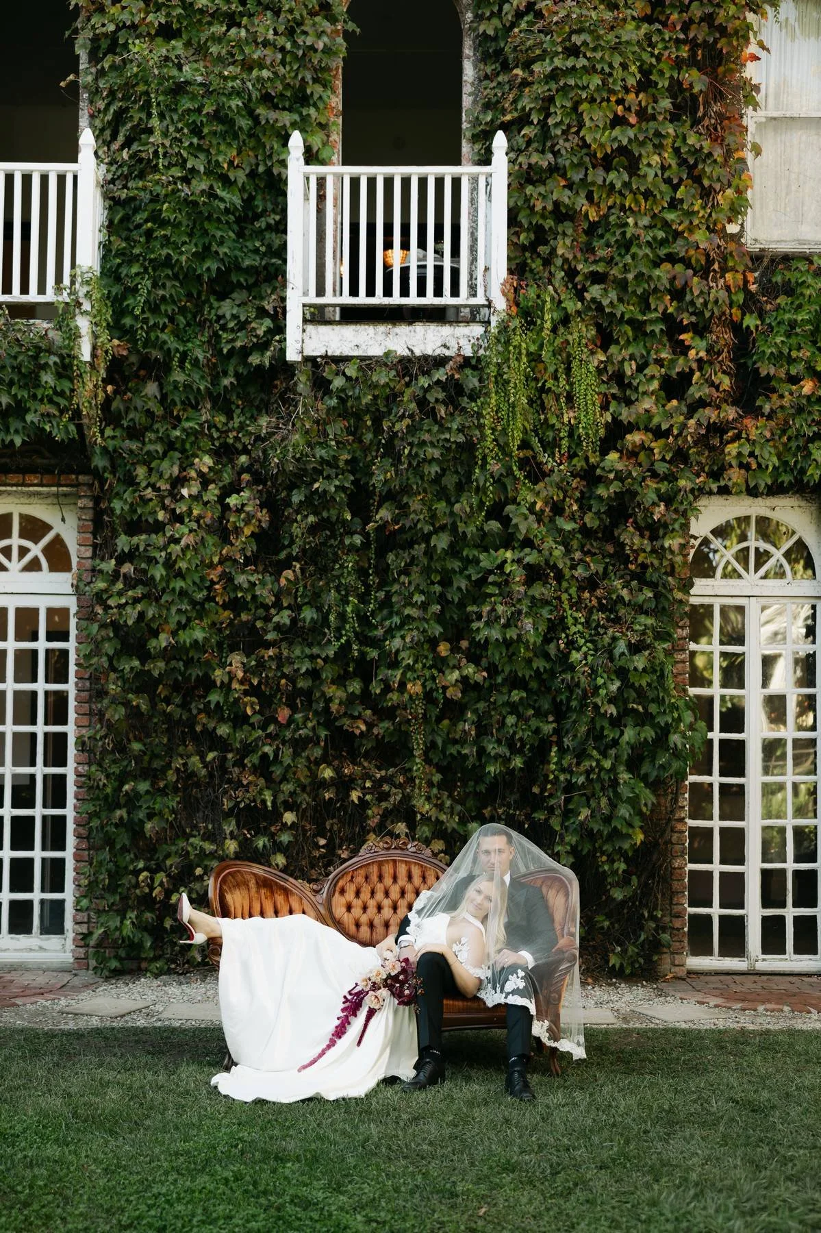 A bride and groom sit on a vintage couch outdoors, partially covered by a wedding veil, in front of a large green ivy-covered building with tall windows and a small balcony.